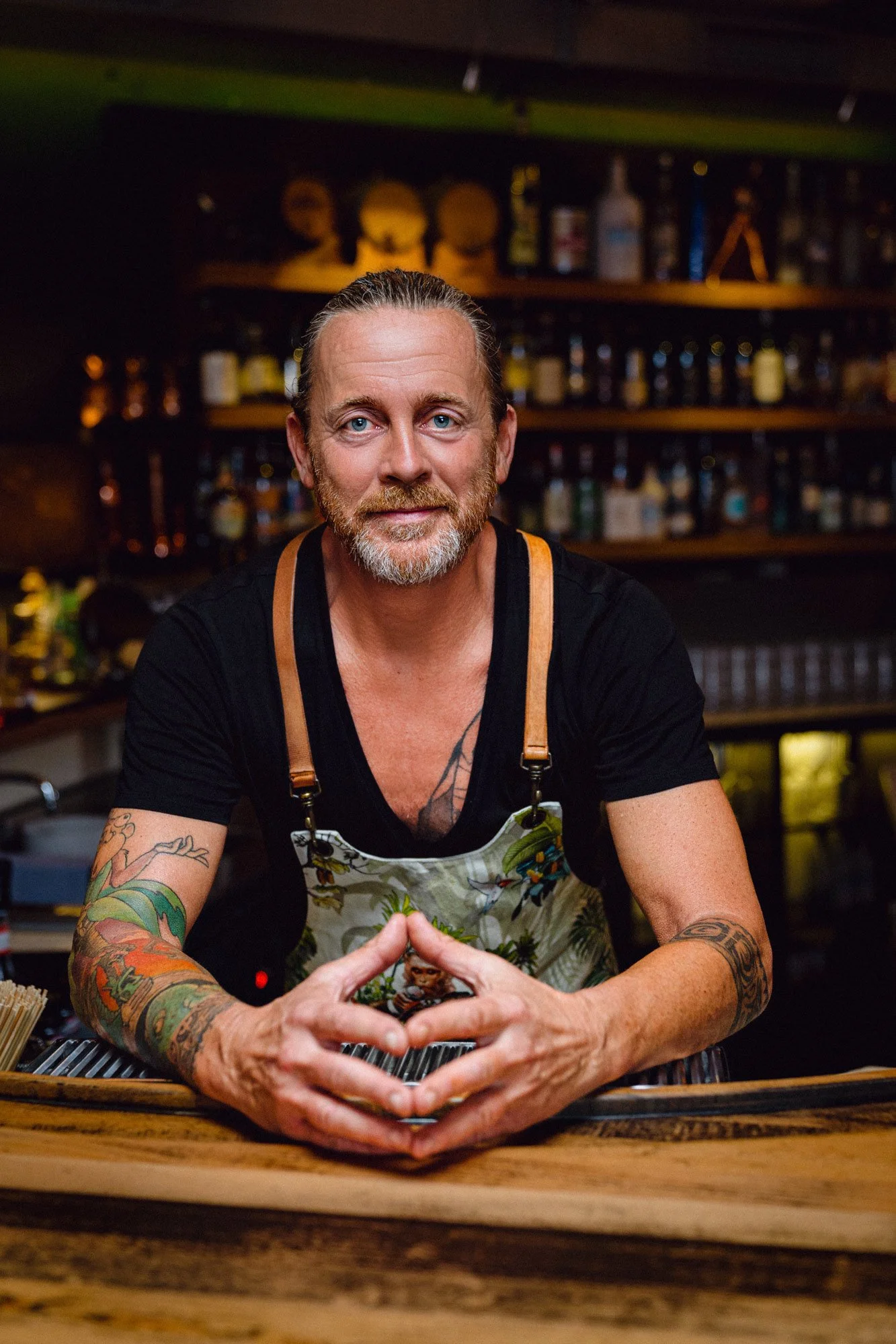 Ein Mann mit Tattoos trägt ein schwarzes T-Shirt und eine bunte Schürze, sitzt an einer Bar, im Hintergrund sind Getränke und Barutensilien sichtbar.