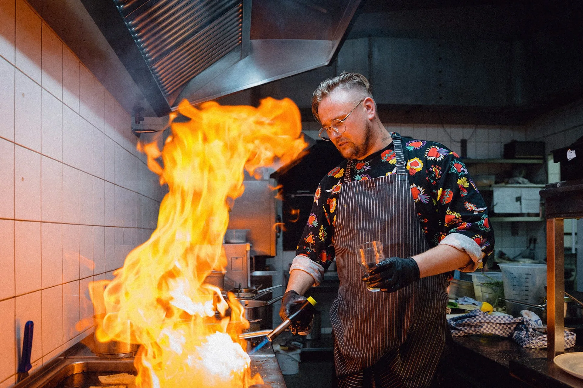 Koch in einer schwarzen Schürze mit bunten Mustern, der Flammen in der Küche erzeugt, während er mit einem Feuerlöscher arbeitet.