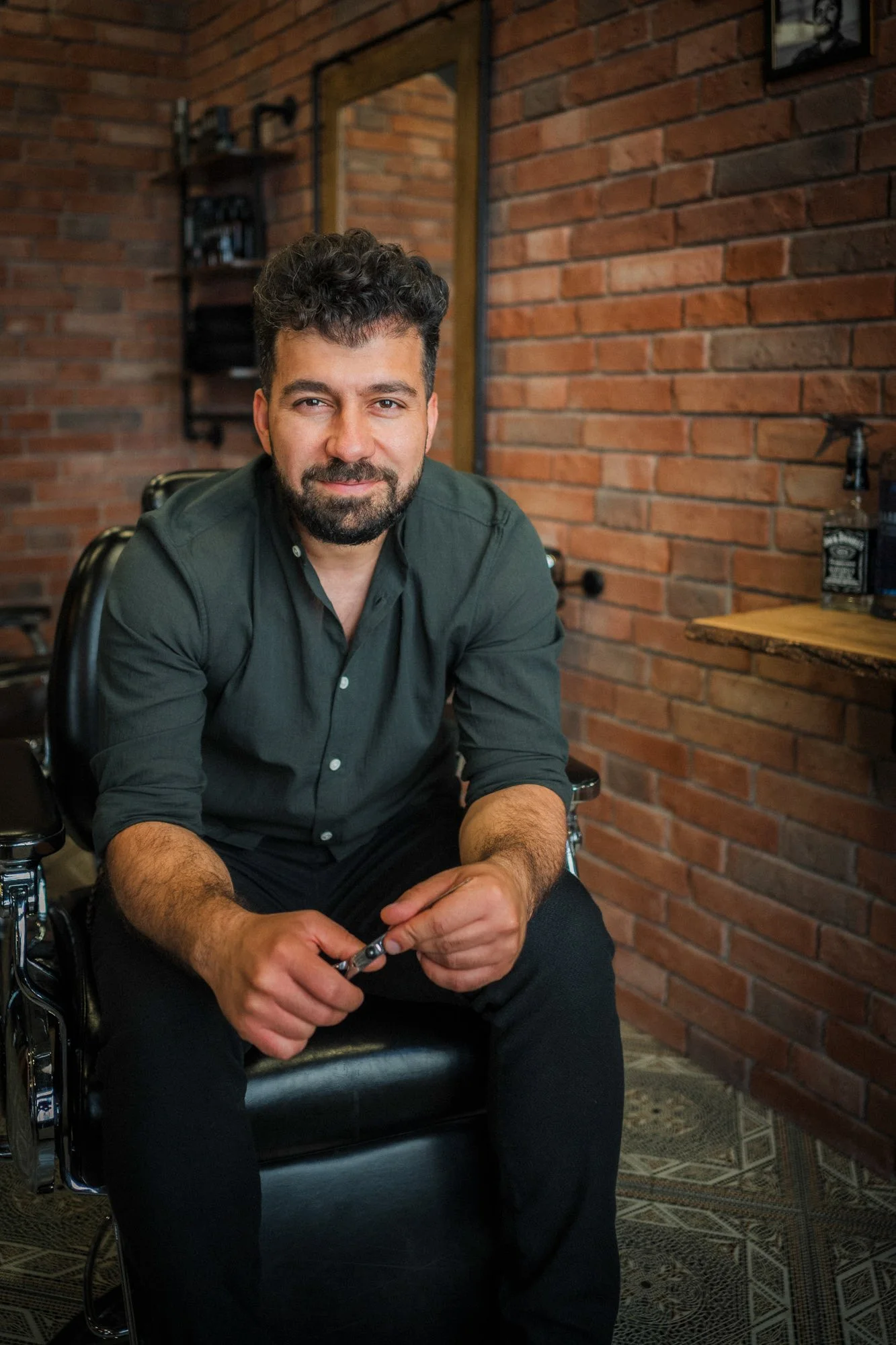 Ein Mann mit dunklem, lockigem Haar und Bart sitzt in einem Friseursalon auf einem schwarzen Friseurstuhl, hält einen Rasiermesser und lächelt leicht. Im Hintergrund sind eine Wand mit Spiegel und Regalen sowie eine Wand im Retro-Stil zu sehen.