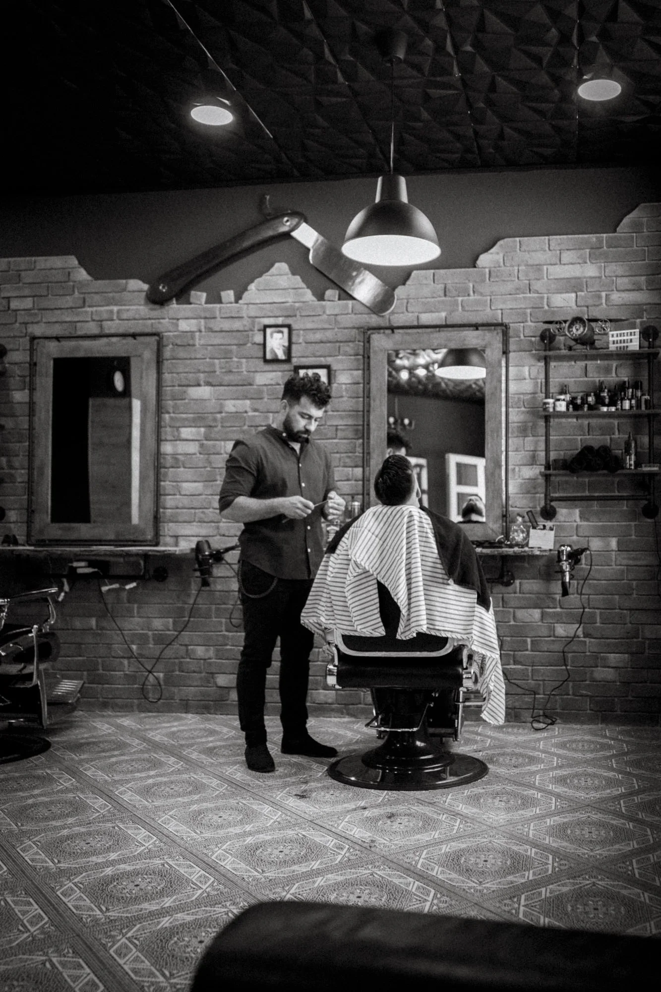 Ein Friseursalon mit einem Kunden, der im Stuhl sitzt und von einem Friseur behandelt wird. Der Friseur steht neben ihm und hält eine Schere. Der Raum ist mit Ziegelwänden, Spiegeln und Regalen mit Haarpflegeprodukten eingerichtet, alles in Schwarz-Weiß.