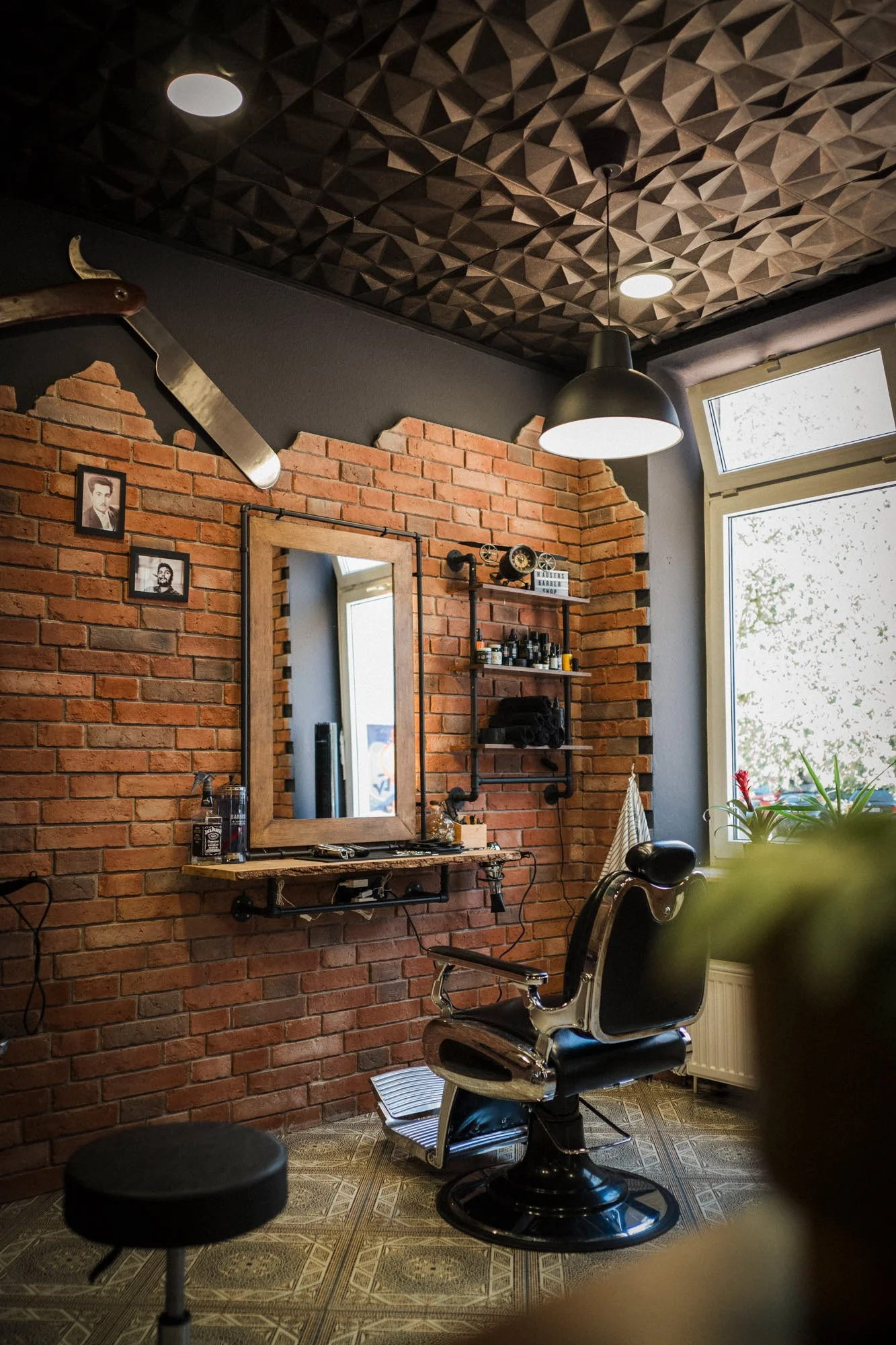 Saal im Friseursalon mit einem schwarzen traditionellen Friseurstuhl, einem Regal mit Haarpflegeprodukten, einem großen Fenster, einer großen Lampe, einer Spiegelwand vor einer roten Backsteinmauer, Fotografien an der Wand und einem Pflanzenn.