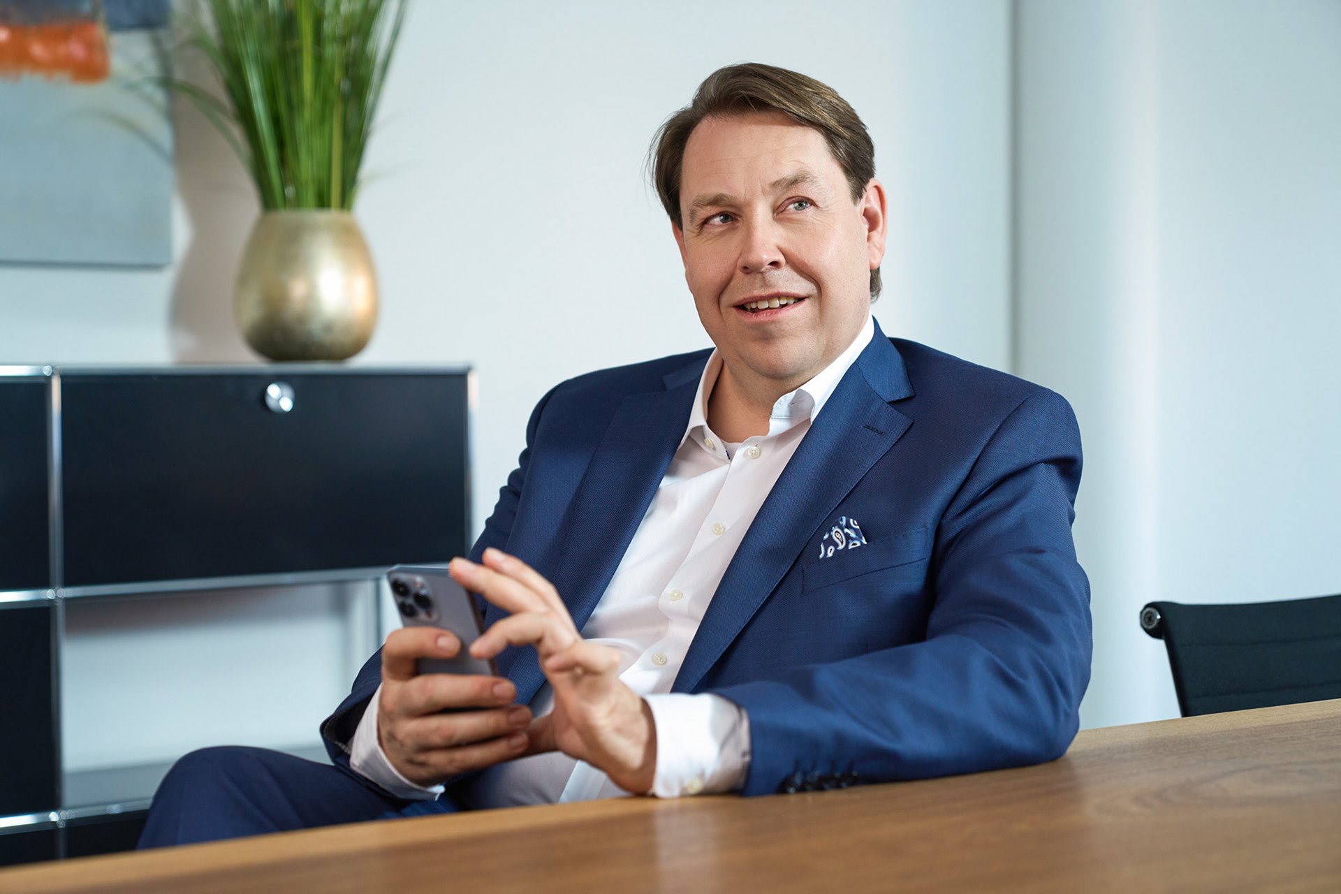 Ein Mann in einem blauen Anzug sitzt an einem Tisch und hält ein Smartphone in der Hand, lächelt und schaut leicht nach oben.