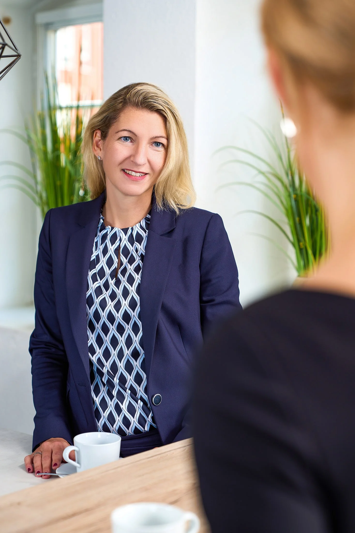Zwei Frauen im Gespräch in einem hellen, modernen Raum mit Pflanzen, eine trägt eine dunkelblaue Jacke und eine blumige Bluse, die andere trägt Schwarz, eine Tasse Kaffee auf dem Tisch.