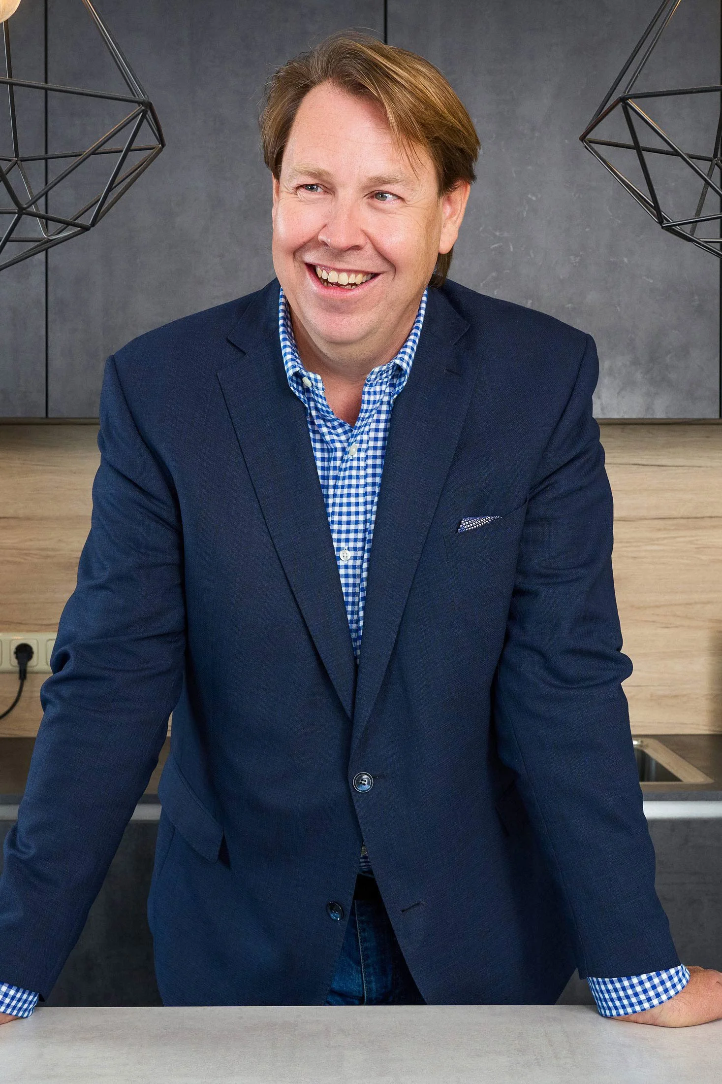 Lächelnder Mann in blauer Jacke und kariertem Hemd, hinter einer Küchenarbeitsplatte, mit geometrischen Lampen im Hintergrund.