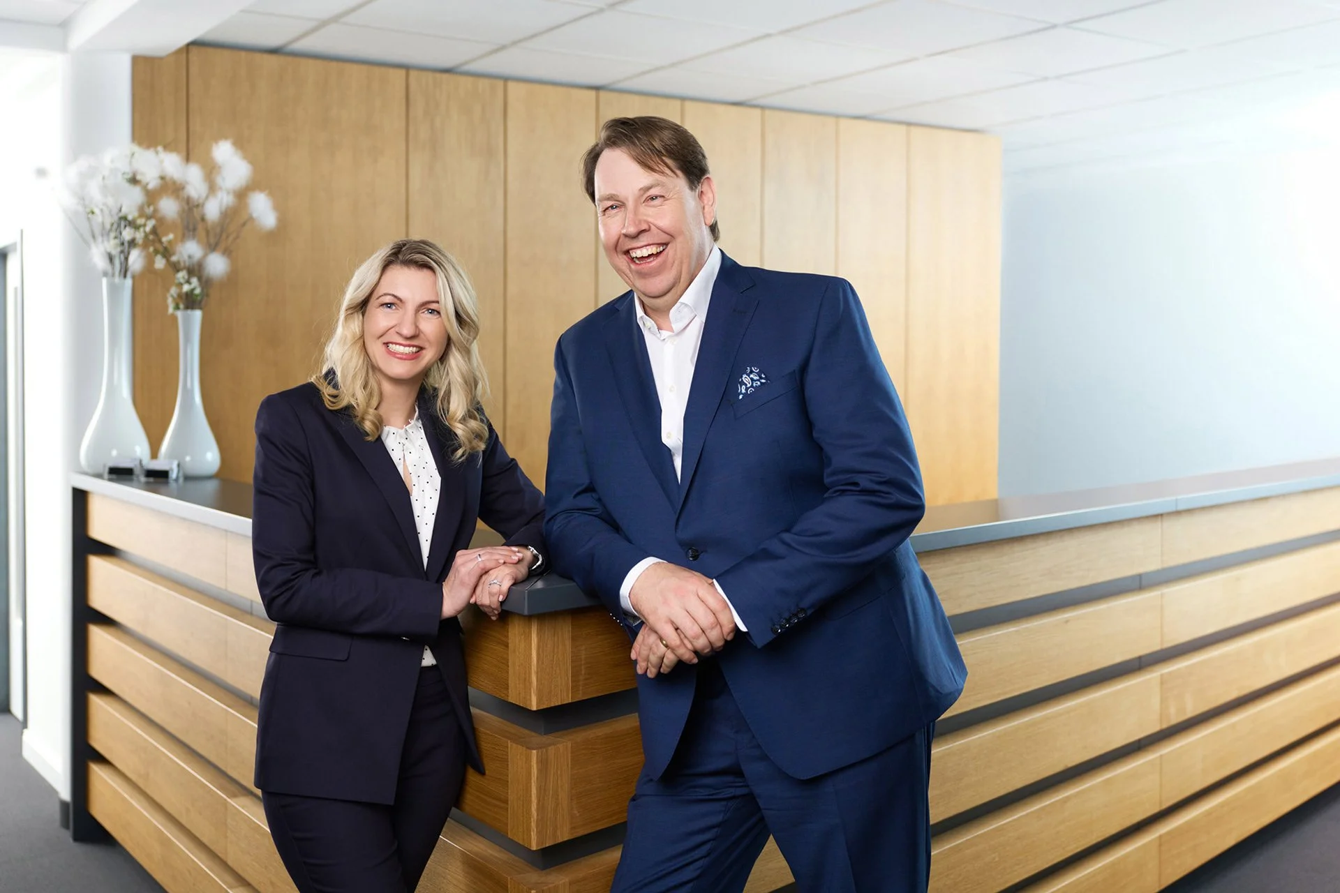 Zwei Geschäftsleute, eine blonde Frau und ein braunhaariger Mann in blauen Anzügen, lachen und posieren an einer Rezeption in einem modernen Büro, mit Holzdekor und weißen Vasen im Hintergrund.