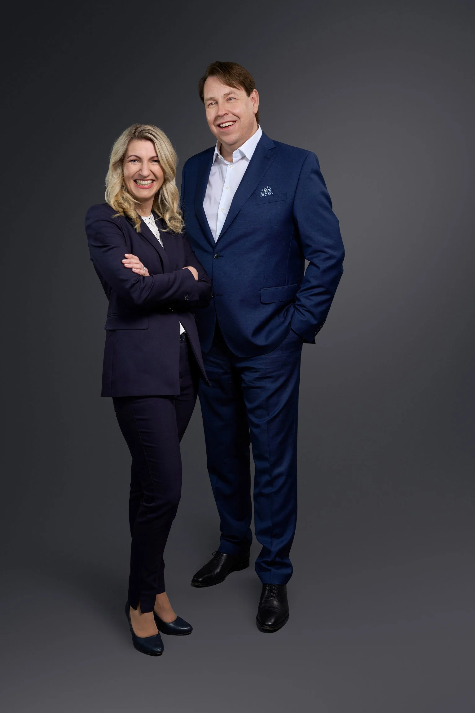 Ein Mann und eine Frau in formeller Kleidung stehen vor einem grauen Hintergrund, lächeln und posieren für die Kamera.