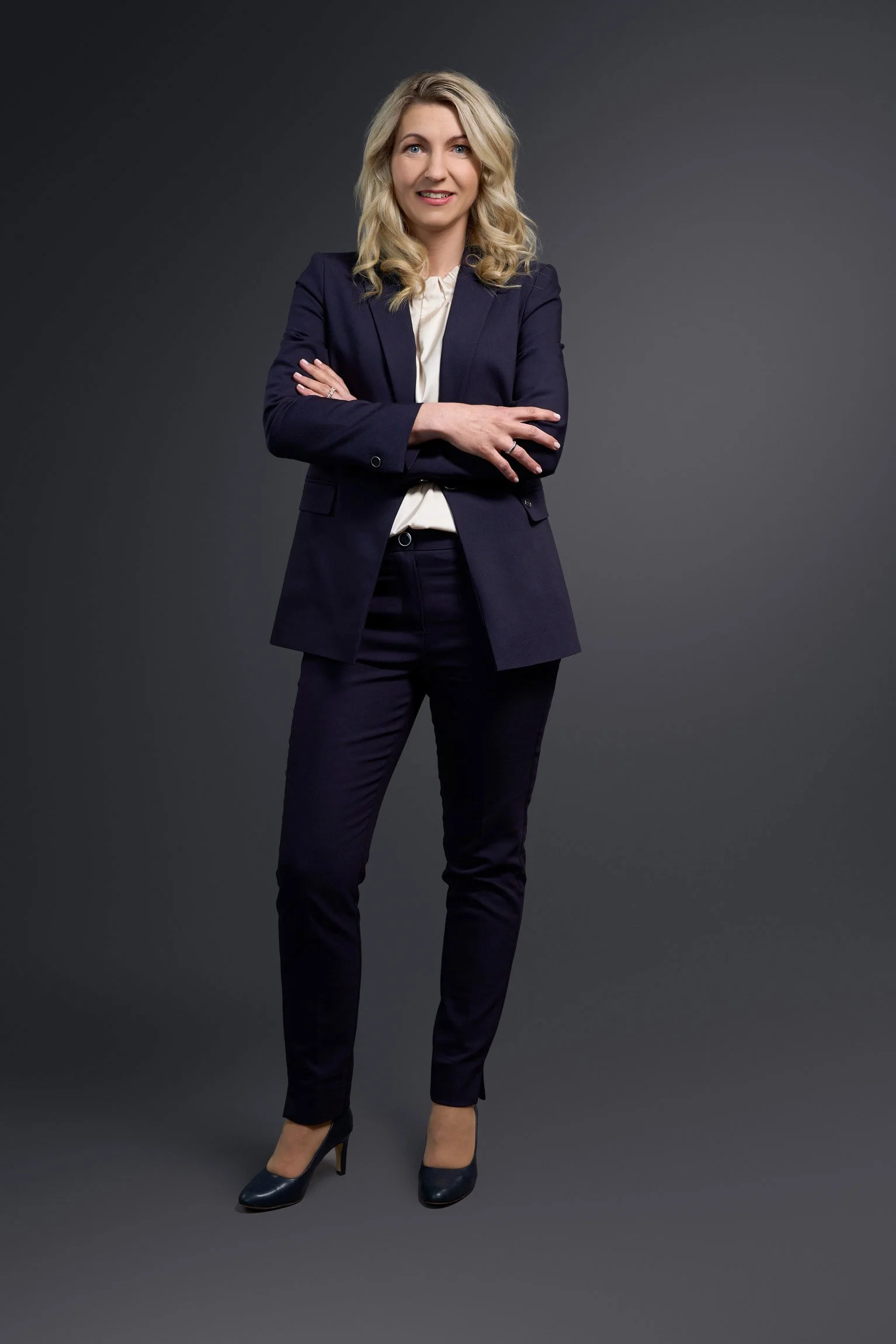Eine Frau in dunkler Businesskleidung steht vor einem grauen Hintergrund.