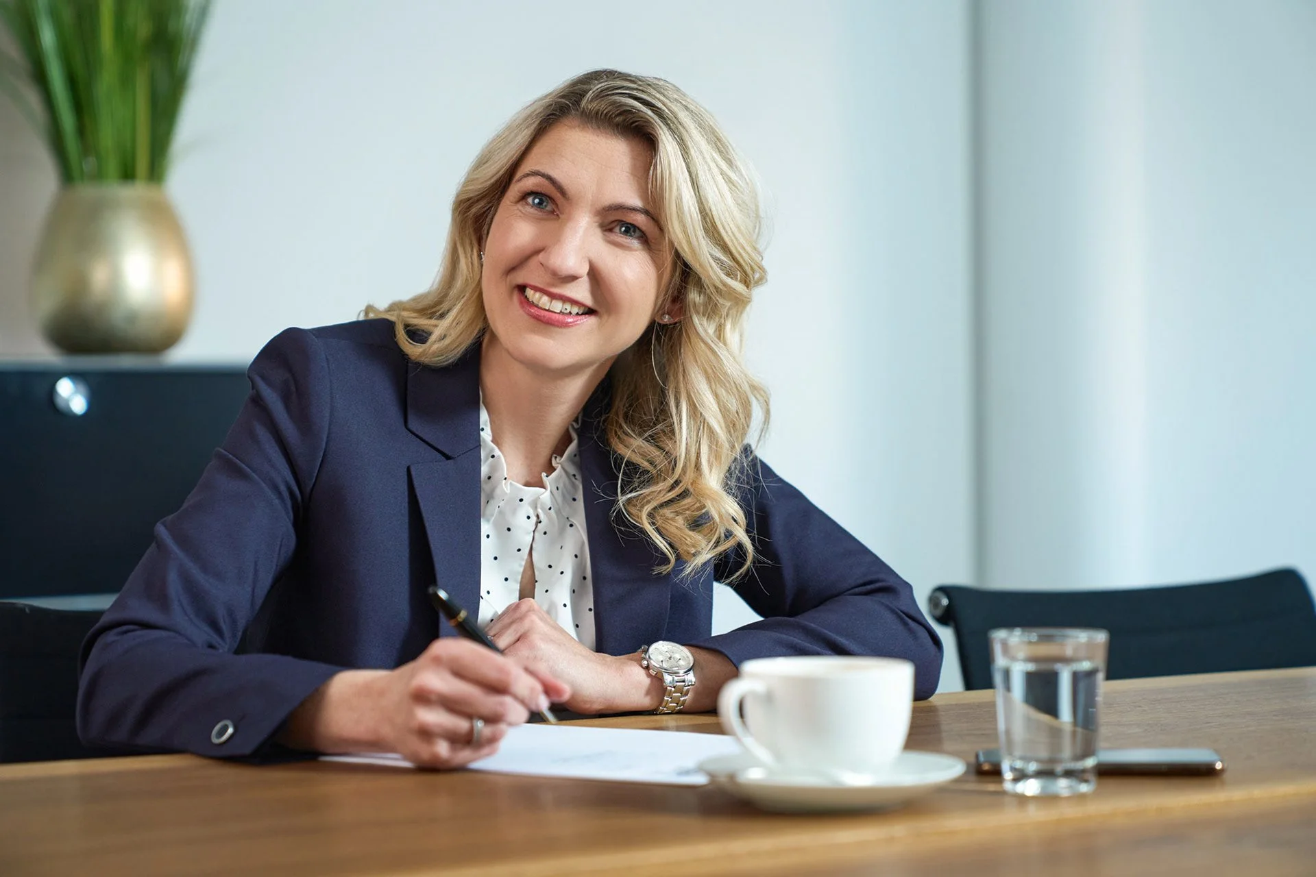 Eine lächelnde Frau im Büro, die an einem Tisch sitzt, mit einer Tasse Kaffee und einem Glas Wasser vor ihr, während sie schreibt.