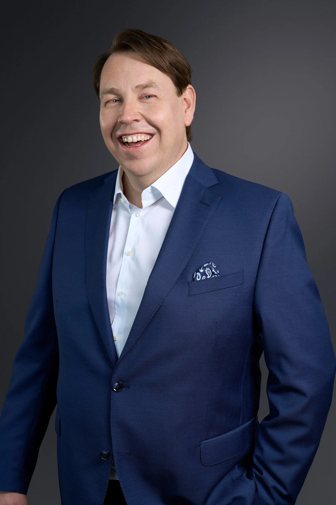 Lächelnder Mann in dunklem blauen Anzug und weißem Hemd vor dunklem Hintergrund.