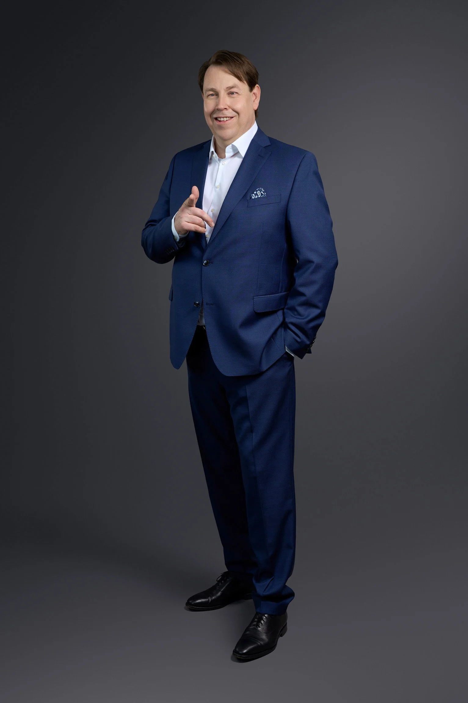 Ein Mann in einem dunkelblauen Anzug steht vor einem grauen Hintergrund und lächelt, während er mit dem Finger in die Kamera zeigt.