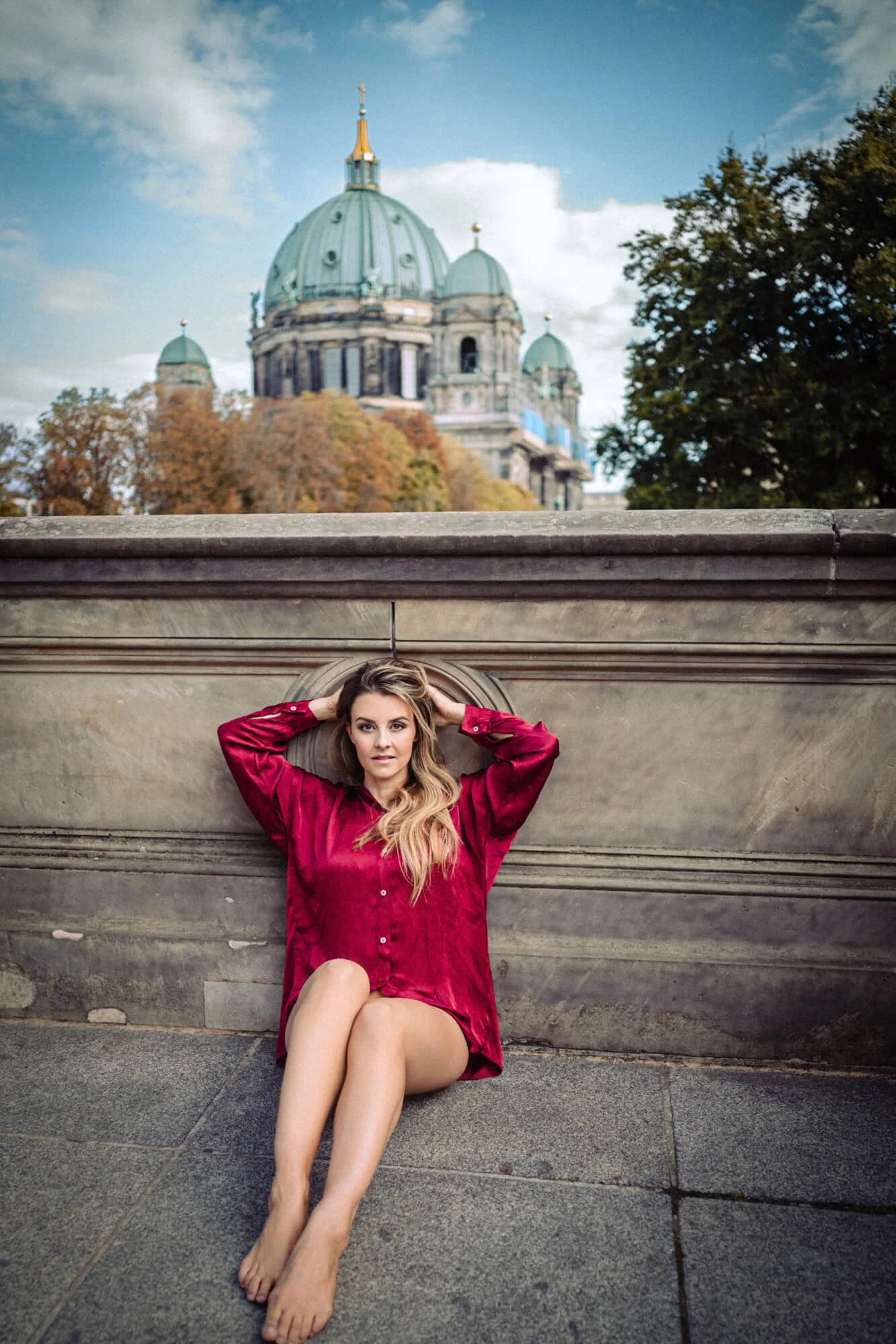 Eine Frau in einem roten Hemd sitzt barfuß auf dem Boden vor einer Steinmauer mit einer großen Kuppel im Hintergrund, vermutlich in Berlin.