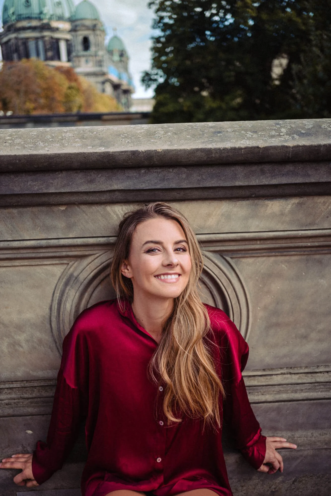 Portrait von Synchronsprecherin Luisa Wietzorek in einer roten Seidenbluse sitzt vor einer Steintreppe, im Hintergrund eine alte Gebäude mit grünen Kuppeln und Bäumen.