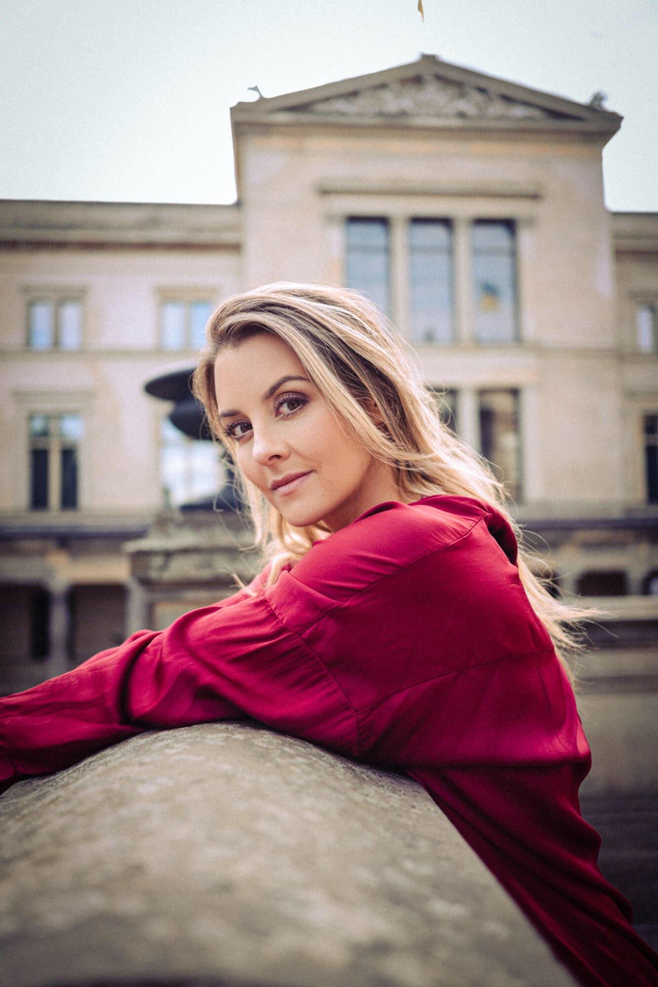 Junge Frau in rotem Hemd liegt auf einer Steinbrüstung vor einem alten Gebäude, blickt in die Kamera, im Hintergrund ein historisches Gebäude mit großen Fenstern.
