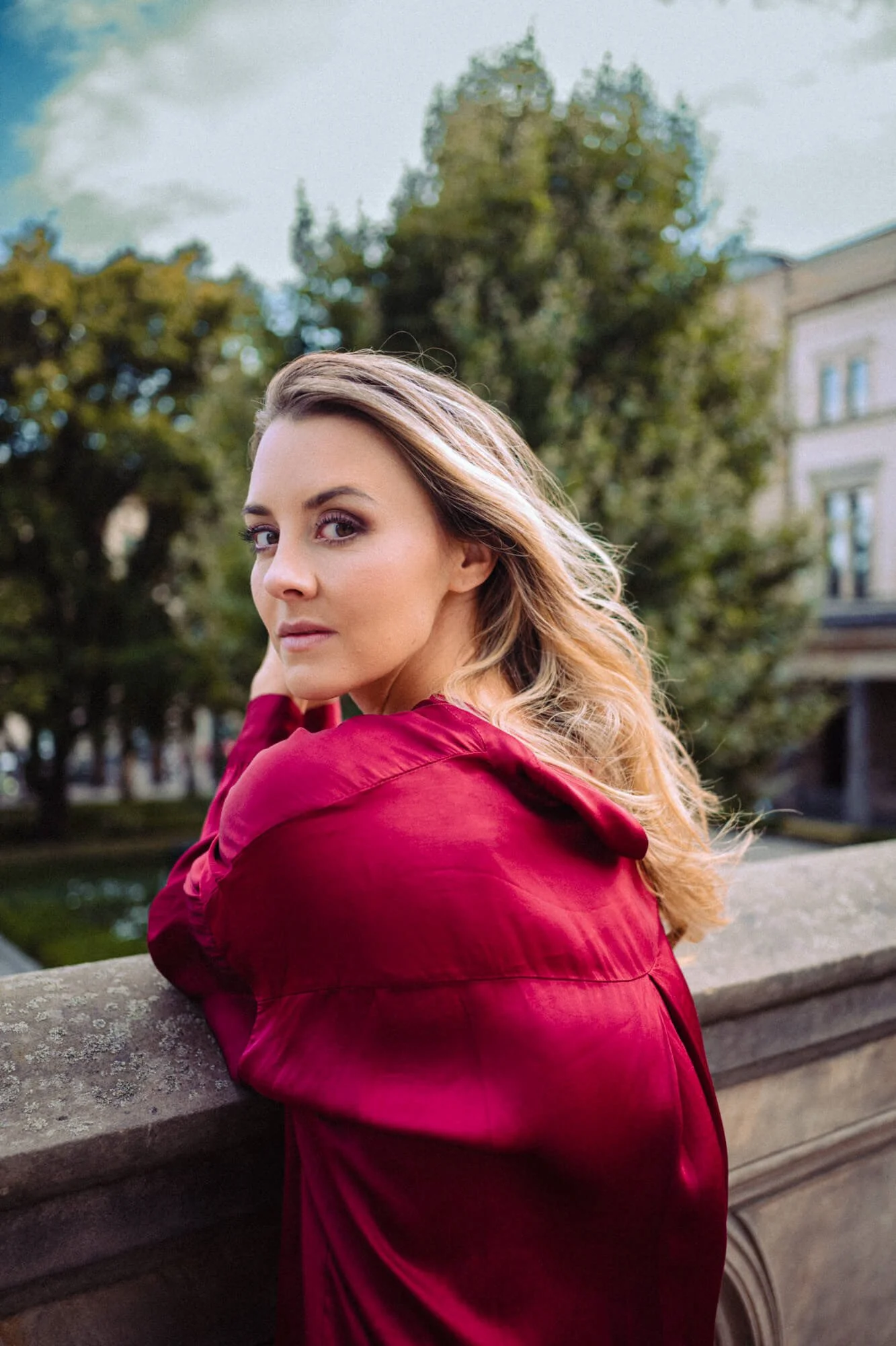 Junge Frau mit blonden, lockigen Haaren, trägt eine rote Jacke und sitzt auf einem Brückengeländer in einem Park mit Bäumen im Hintergrund.