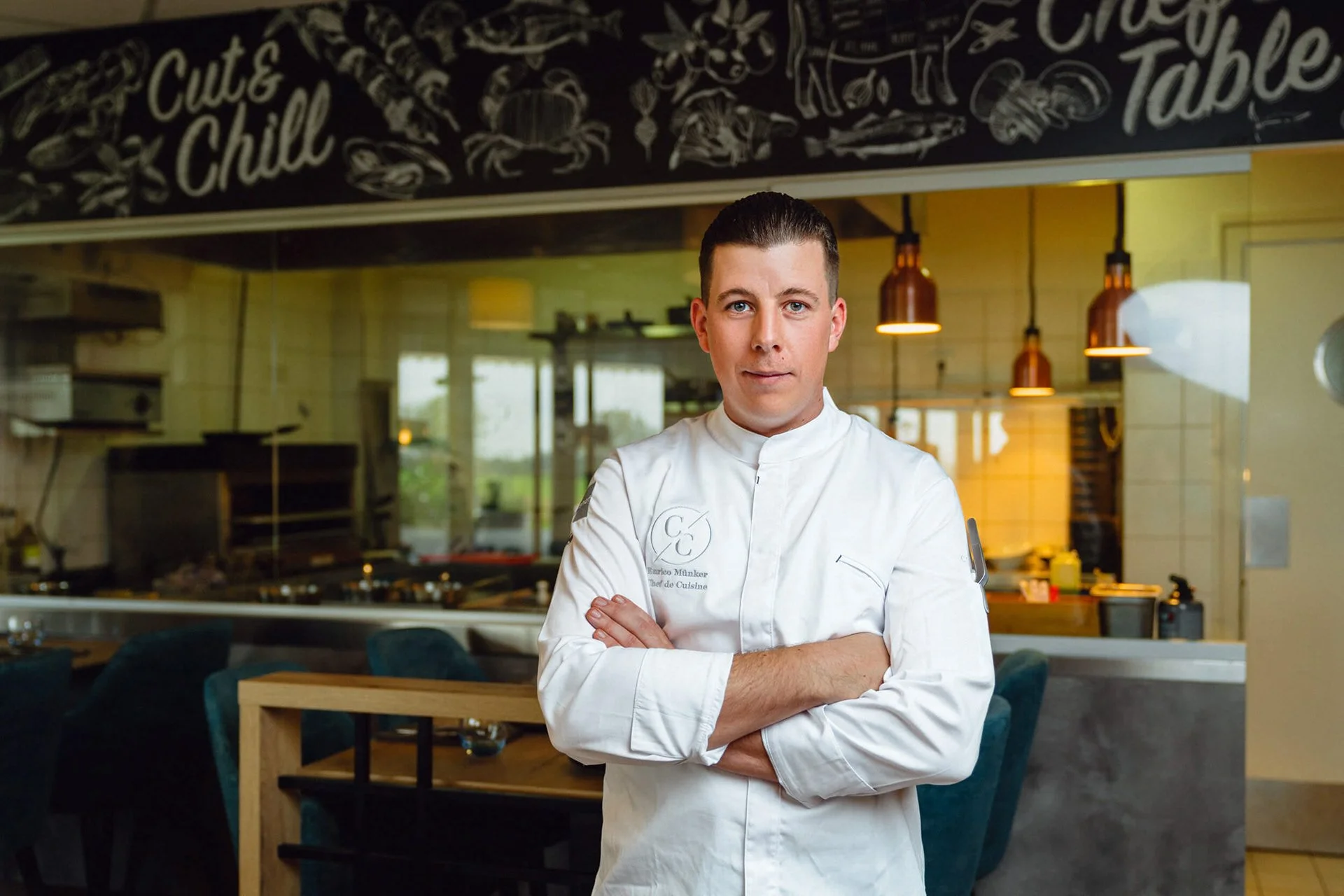 Ein Koch in weißer Uniform steht mit verschränkten Armen in einem Restaurantküchenraum, im Hintergrund sind Tische, Stühle und Küchengeräte sichtbar.