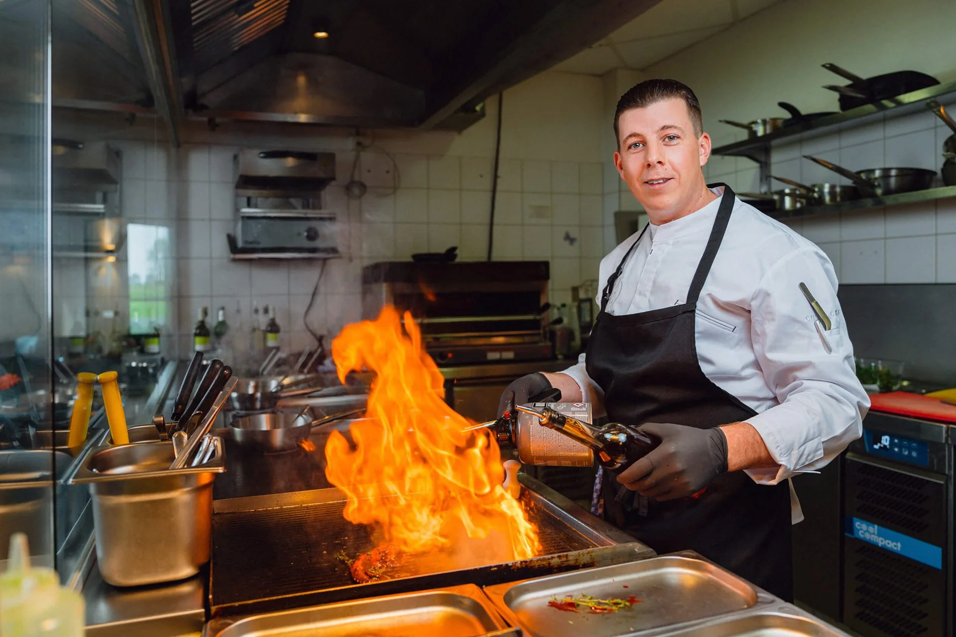 Koch in Küche mit Flamme beim Zubereiten eines Gerichts.