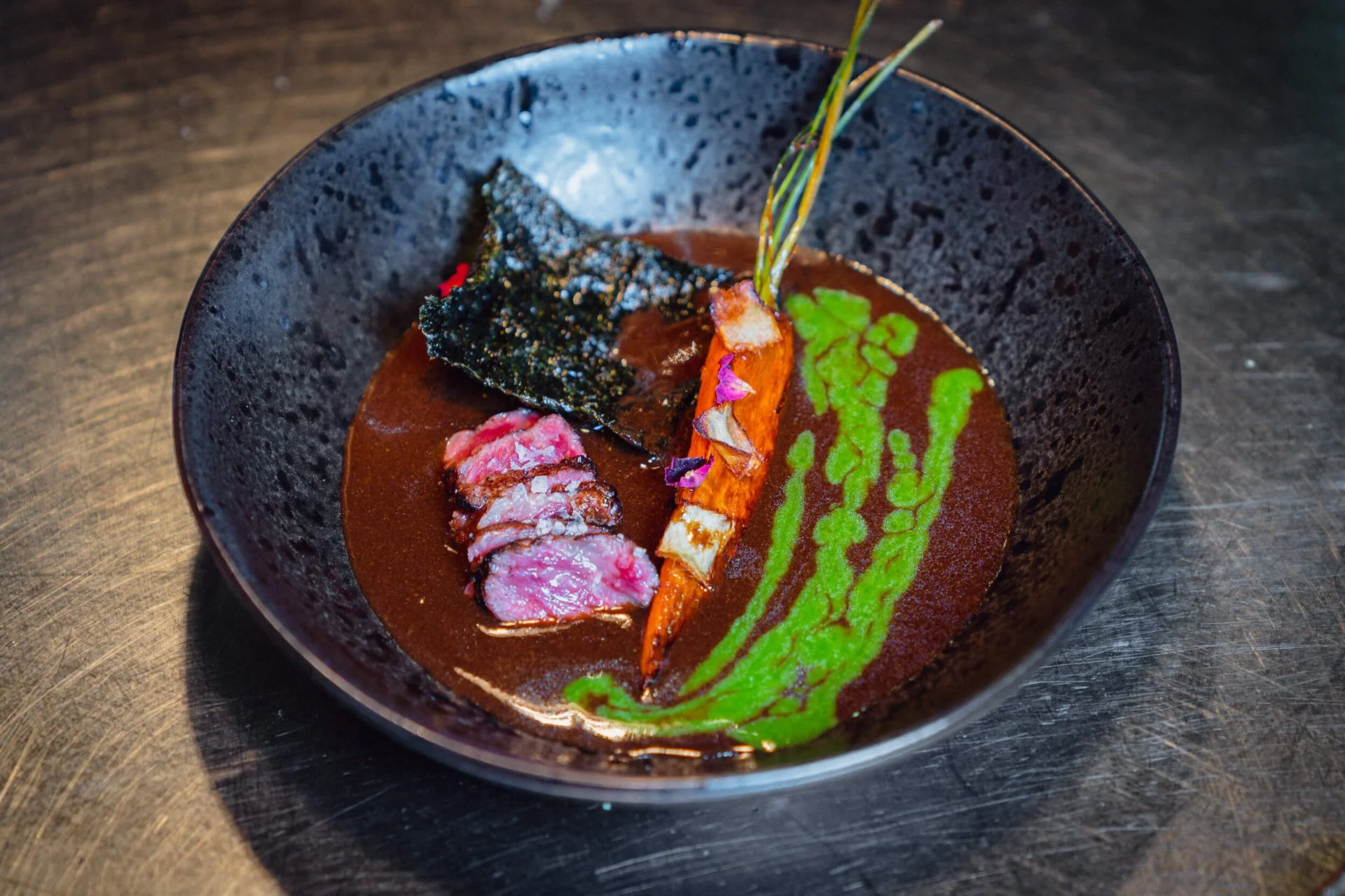 Ein schwarzer Suppenteller mit brauner Suppe, Garnelen, Seetang, Gemüse und einer grünen Soße, dekorativ angerichtet.