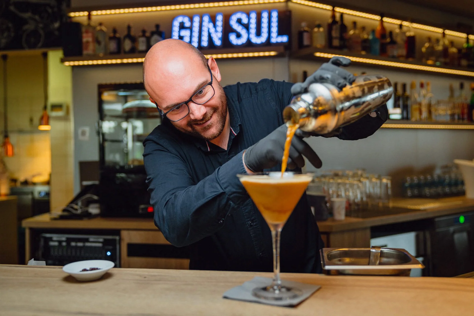 Ein Barkeeper gießt einen Cocktail aus einem Shaker in ein Martiniglas, trägt eine schwarze Schürze und trägt schwarze Handschuhe, im Hintergrund ist ein Bar-Schild mit leuchtendem Schriftzug 'GINSUL' zu sehen.