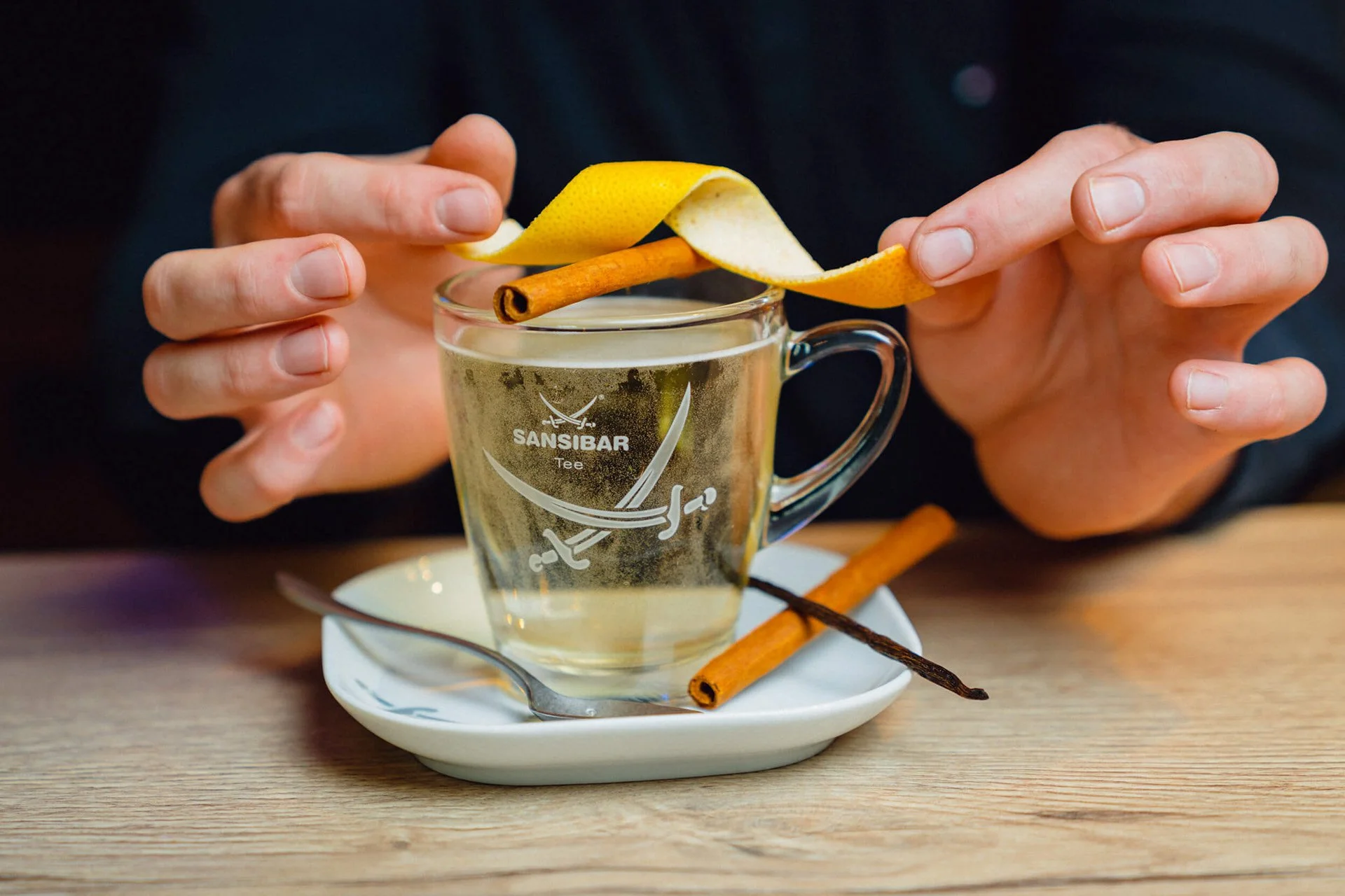 Eine Tasse heißer Tee mit Zitrusschale, Zimtstange und Vanilleschote, auf einem weißen Untersetzer, auf einem Holztisch. Eine Person berührt die Zitrusschale. Das Logo auf der Tasse zeigt gekreuzte Schwerter und den Schriftzug 'SANSIBAR'.