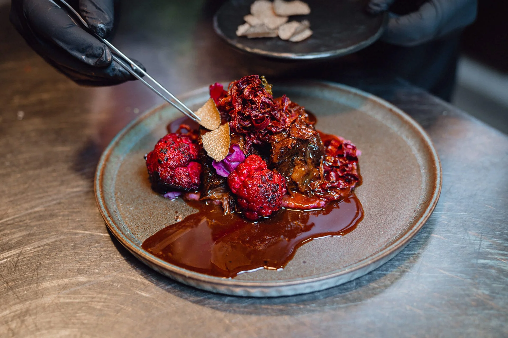 Ein Teller mit aufwändig zubereitetem Gericht, das verschiedene Beeren, Sauce und essbare Blumen enthält, wird von einem Koch mit schwarzen Handschuhen serviert.