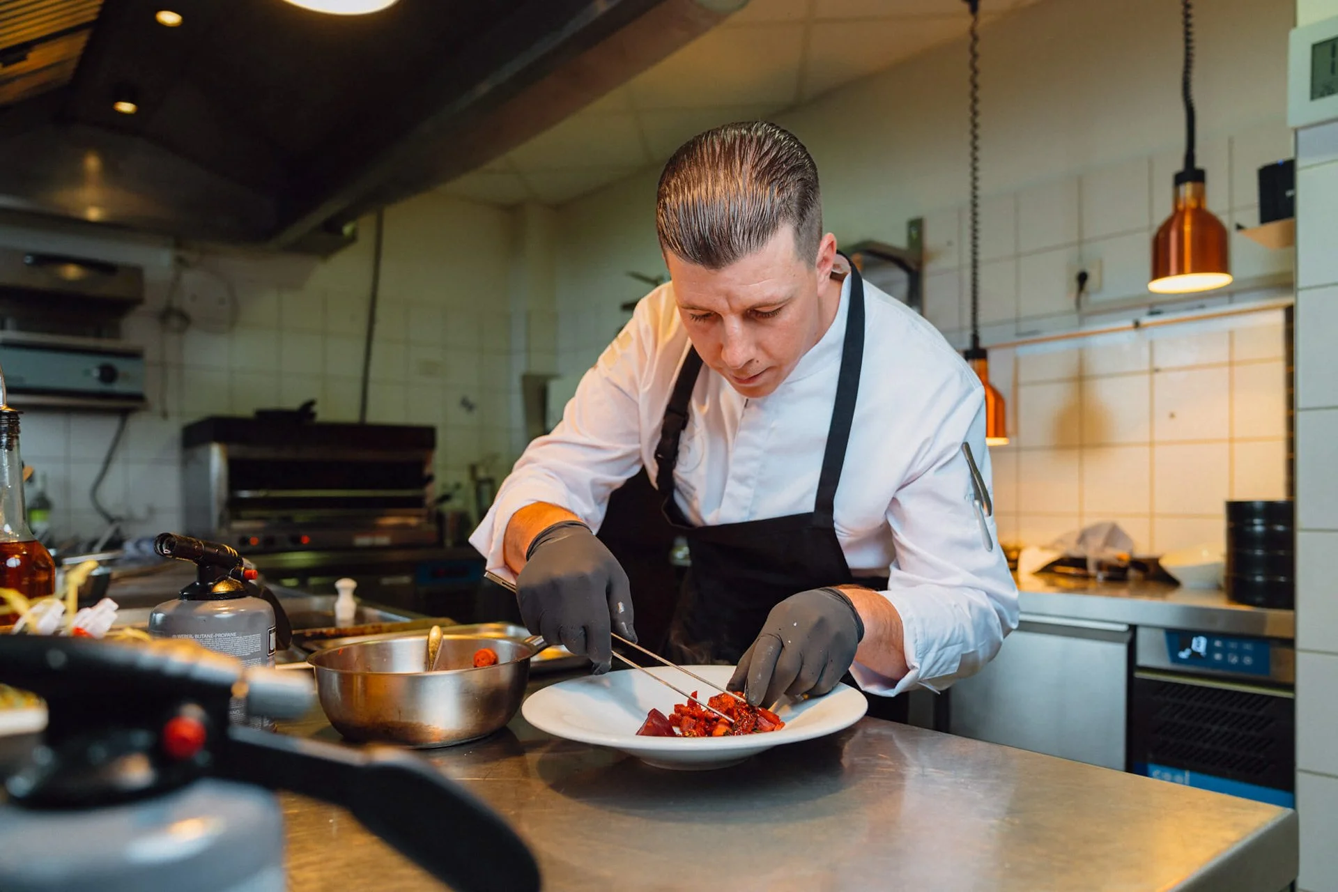 Ein Koch bereitet in einer professionellen Küche ein Gericht zu, trägt schwarze Handschuhe und einen weißen Kochmantel.