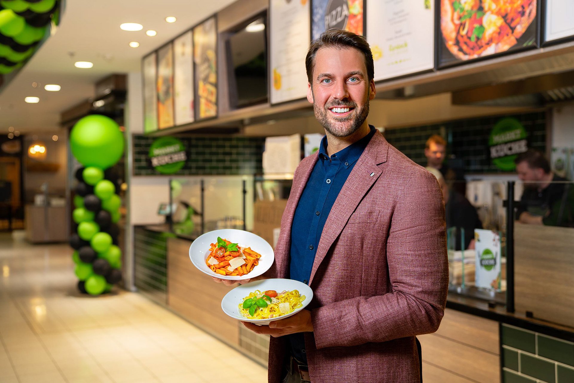 Stephan Cunäus in einem roten Blazer hält zwei Teller mit Pasta in der Marktküche Rostock im Warnowpark.