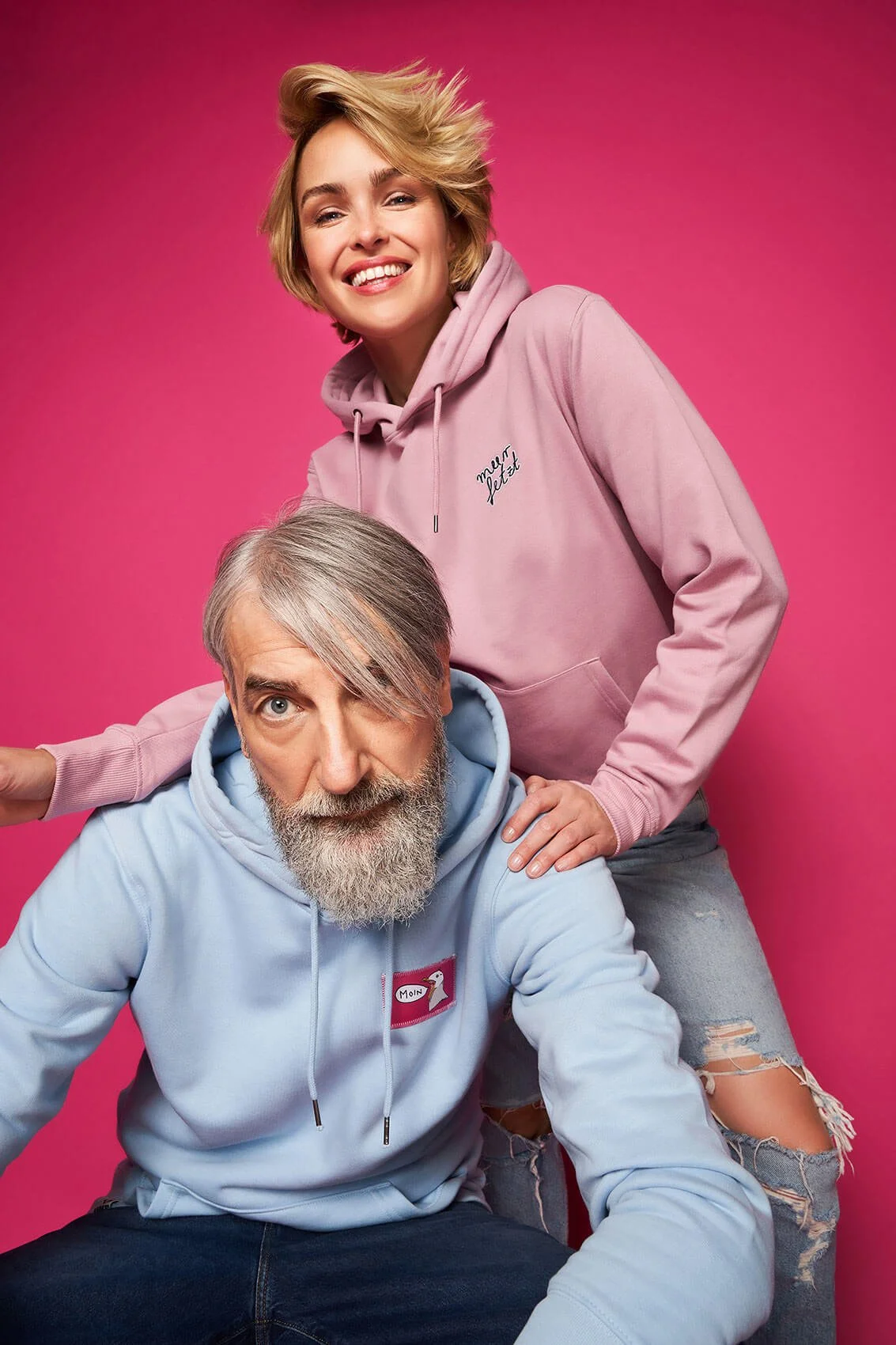 Ein junger Mann mit grauem Bart und einem blauen Hoodie sitzt, während eine junge Frau mit kurzen, lockigen Haaren und rosa Hoodie hinter ihm steht und beide vor einem pinken Hintergrund sind.