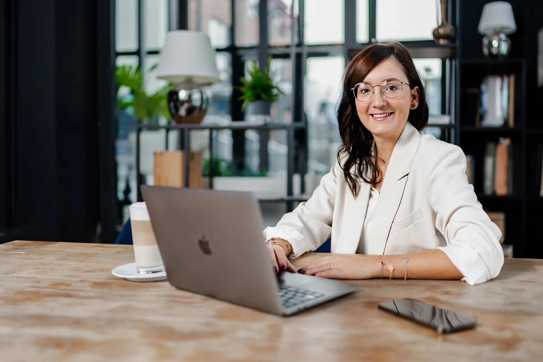 Multiprofessionelle Frau mit Brille und weißen Blazer arbeitet an einem Laptop in einem modernen Büro mit offenem Regal im Hintergrund.