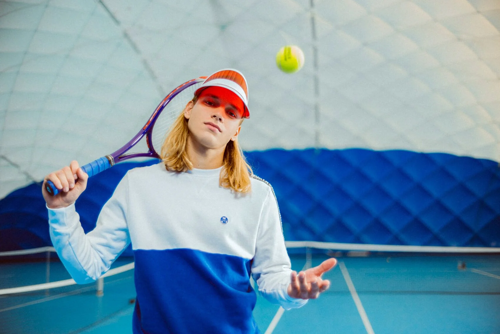 Junge Frau im Tennisraum mit Tennisschläger, Turnschuhen, weiß-blauem Sweatshirt, Pose mit einer Hand, Tennisball in der Luft.