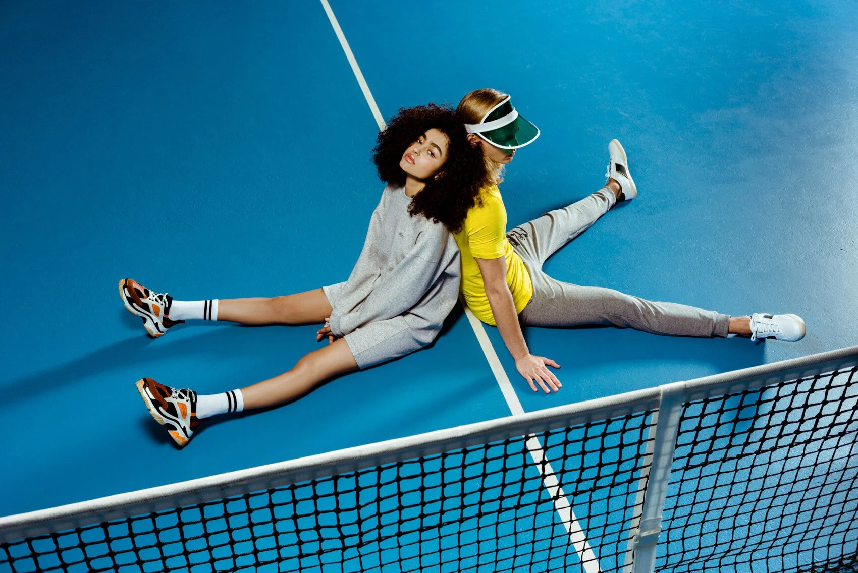 Zwei Frauen liegen rückwärts auf einem blauen Tennisplatz, getrennt durch das Netz. Eine trägt einen grauen Hoodie und Turnschuhe, die andere trägt eine gelbe T-Shirt, graue Hosen und einen Tennisvisier.