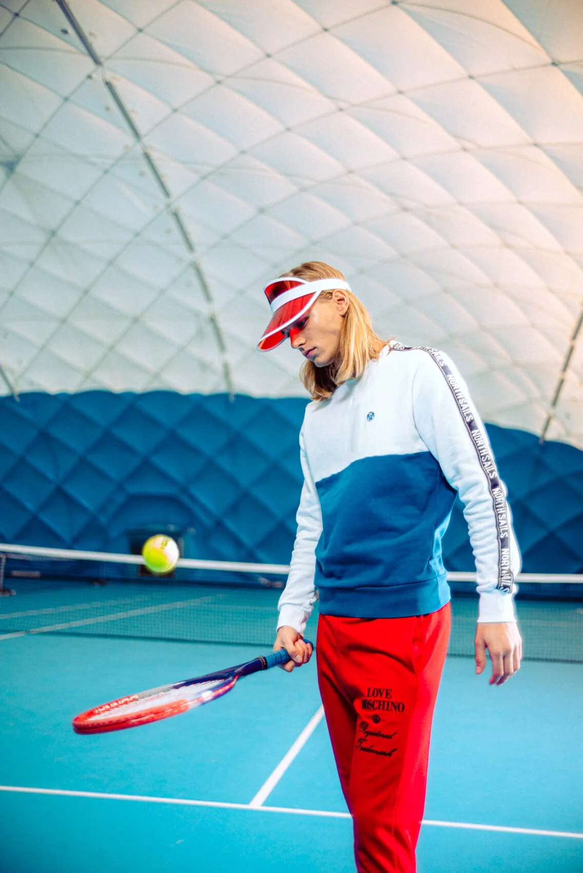 Frau in einem Tennisclub beim Training, trägt eine weiße und blaue Sweatjacke, rote Turnhose mit Markenlogo, eine rote Visier und Schutzbrille, hält einen Tennisschläger, im Hintergrund ein Tennisball auf dem Platz.