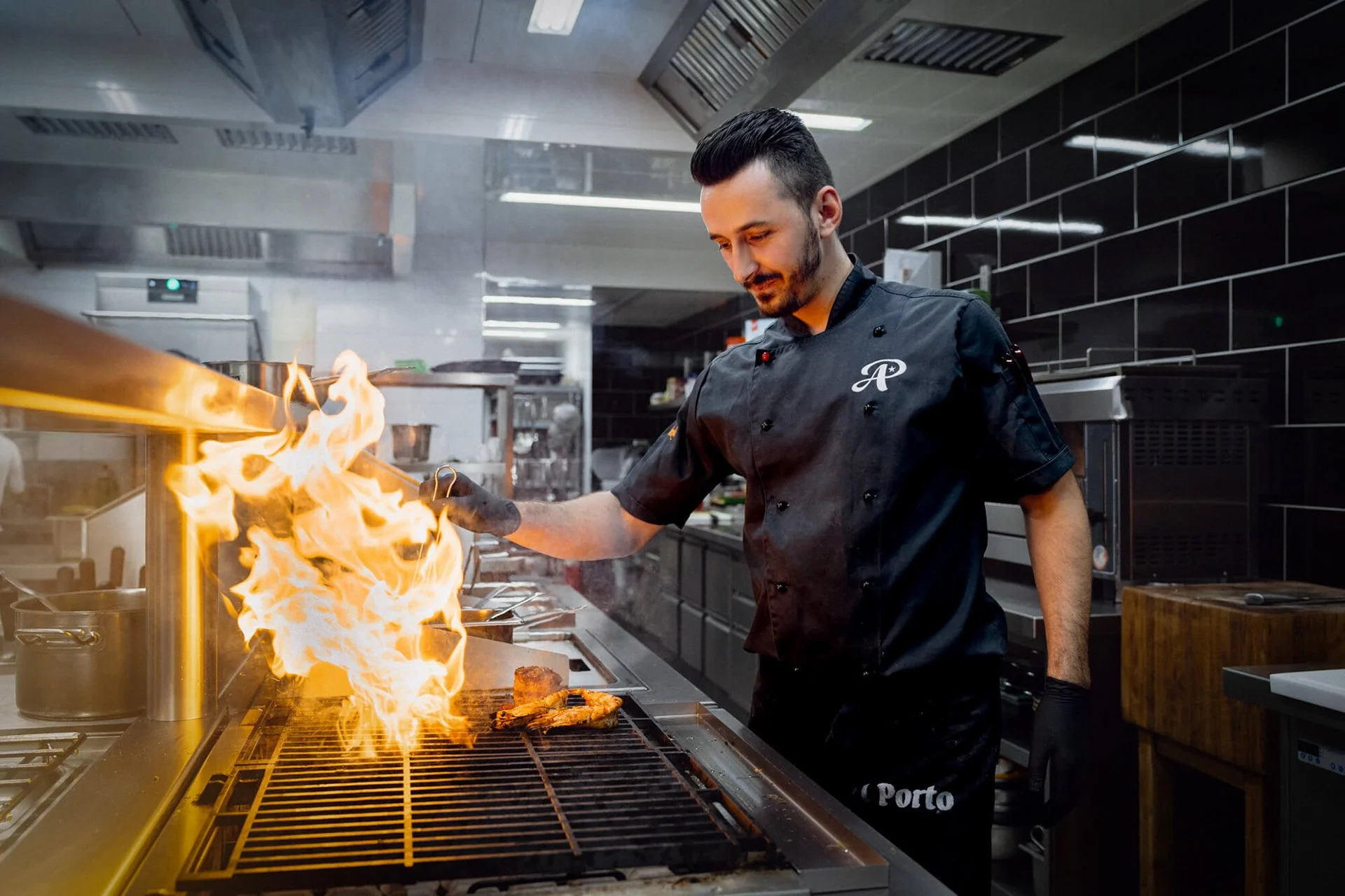 Ein Koch in einem Schwarzkochoutfit steht in einer professionellen Küche und benutzt eine Zange, um Lebensmittel über einer Flamme auf einem Grill zu grillen.