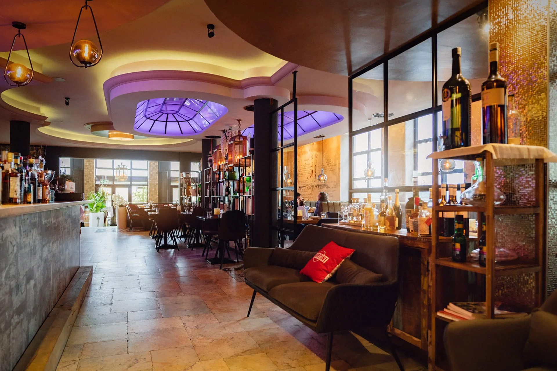 Innenansicht eines modernen Restaurants mit Bar, beleuchteten Deckenlampen und großen Fenstern, gemütliche Sitzgelegenheiten und dekorative Regale mit Flaschen.