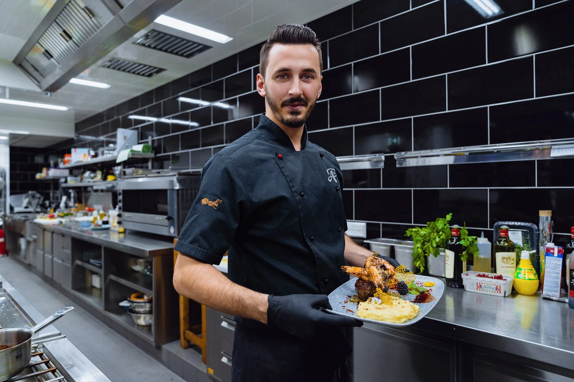 Ein Koch präsentiert ein Tellergericht mit Fleisch, Beilagen und Garnierungen in einer modernen Küche.