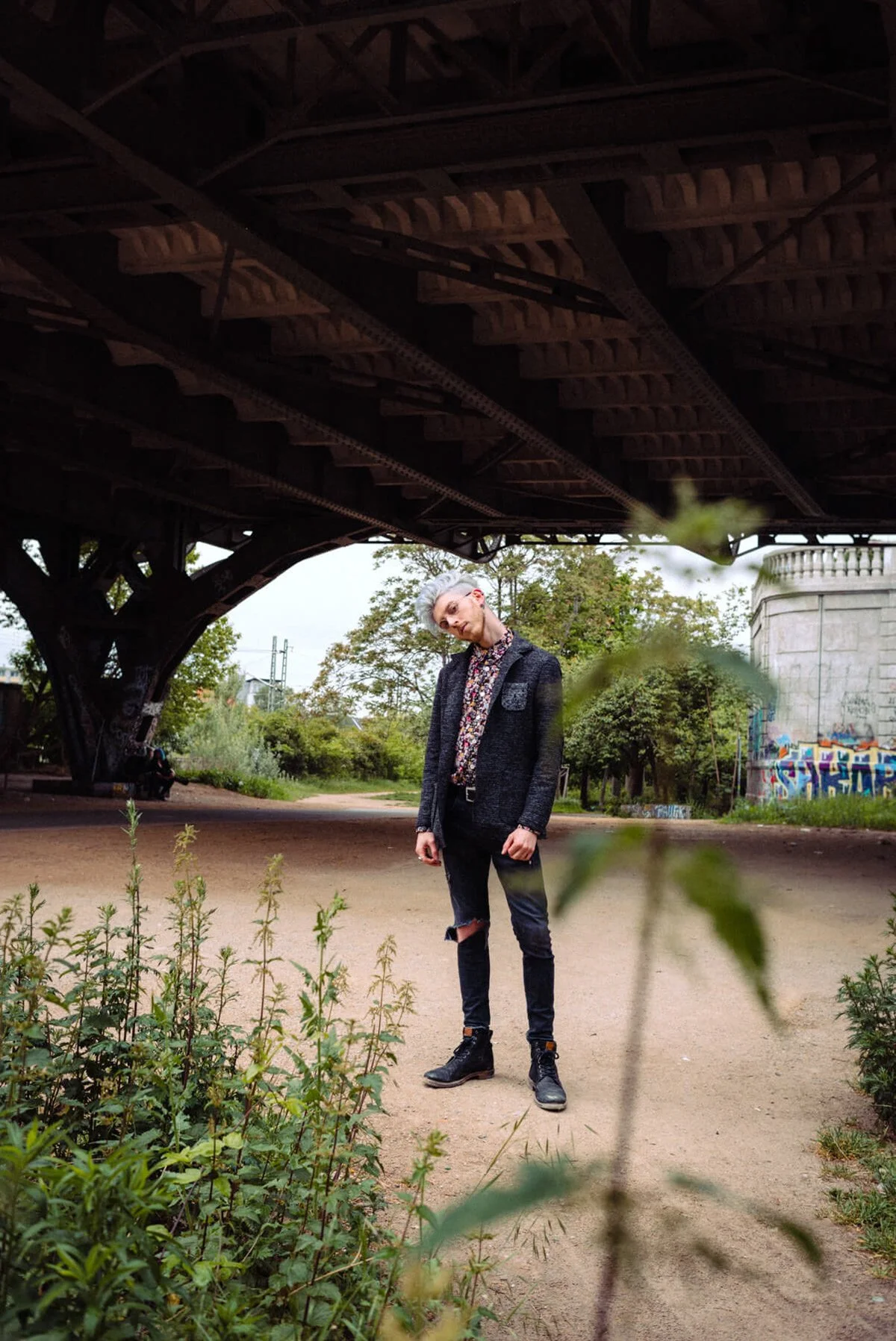 Junge Frau mit kurzen grauen Haaren, dunklem Jeans, einer schwarzen Jacke und Stiefeln steht unter einer Brücke im Freien, umgeben von Büschen und Bäumen.