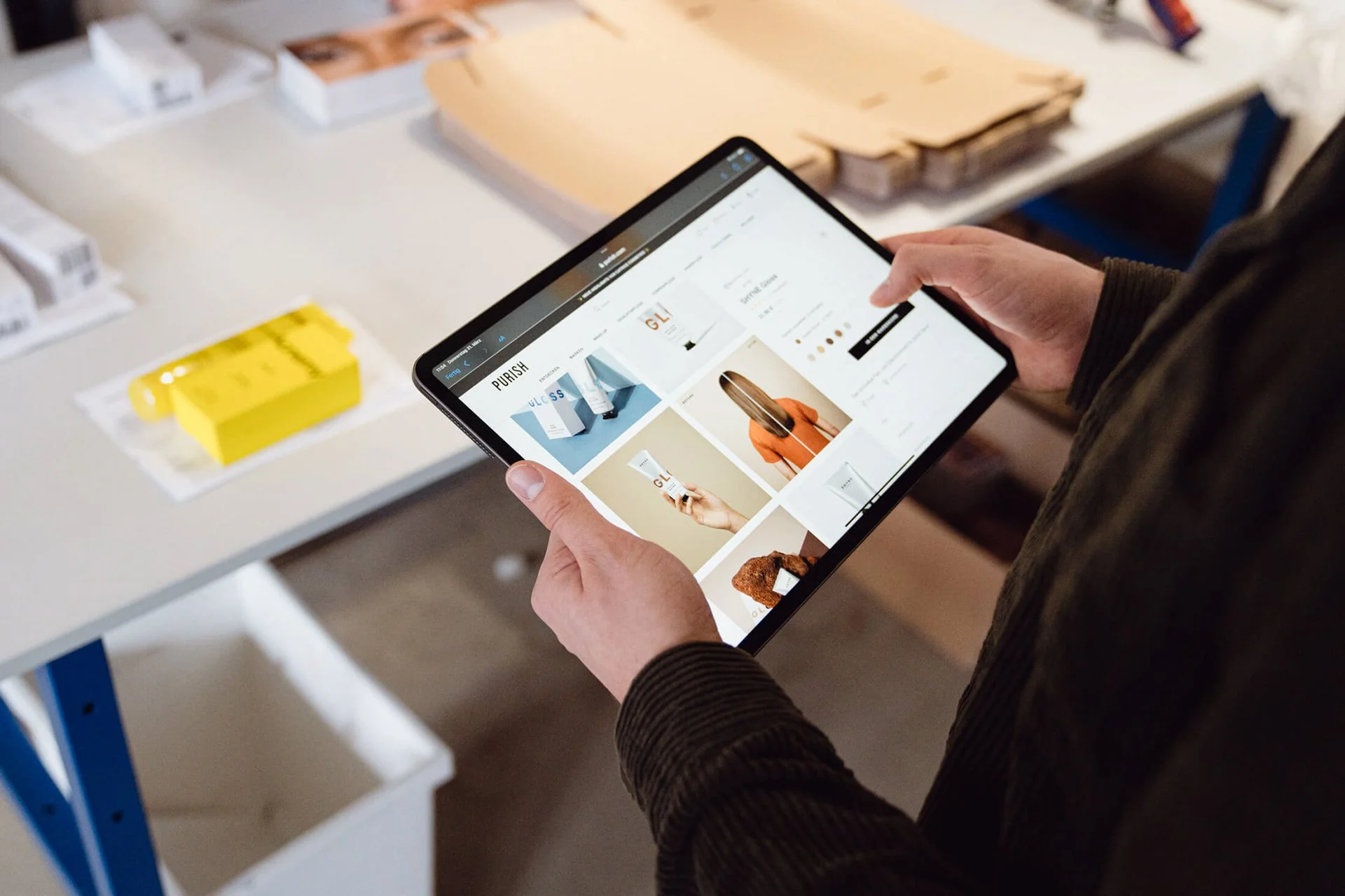 Eine Person hält ein Tablet, auf dem eine Produktseite mit Kosmetikartikeln angezeigt wird. Im Hintergrund sind Verpackungen und Pakete auf einem Tisch sichtbar.