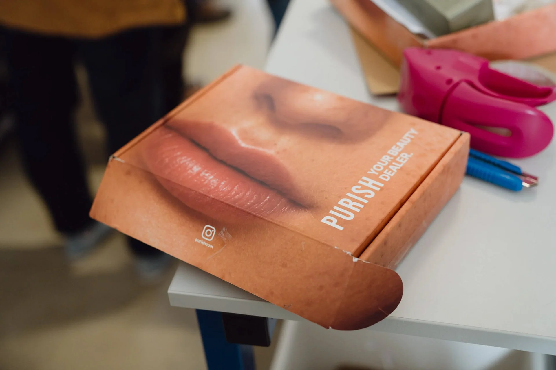 Auf einem weißen Tisch liegt eine bemalte Pappbox mit einem Lippenfoto, darunter steht 'PURISH YOUR BEAUTY' und ein Instagram-Logo. Zusätzlich sind eine pinke Haarschneideschere, ein blauer Kugelschreiber und ein roter Filzstift sichtbar.