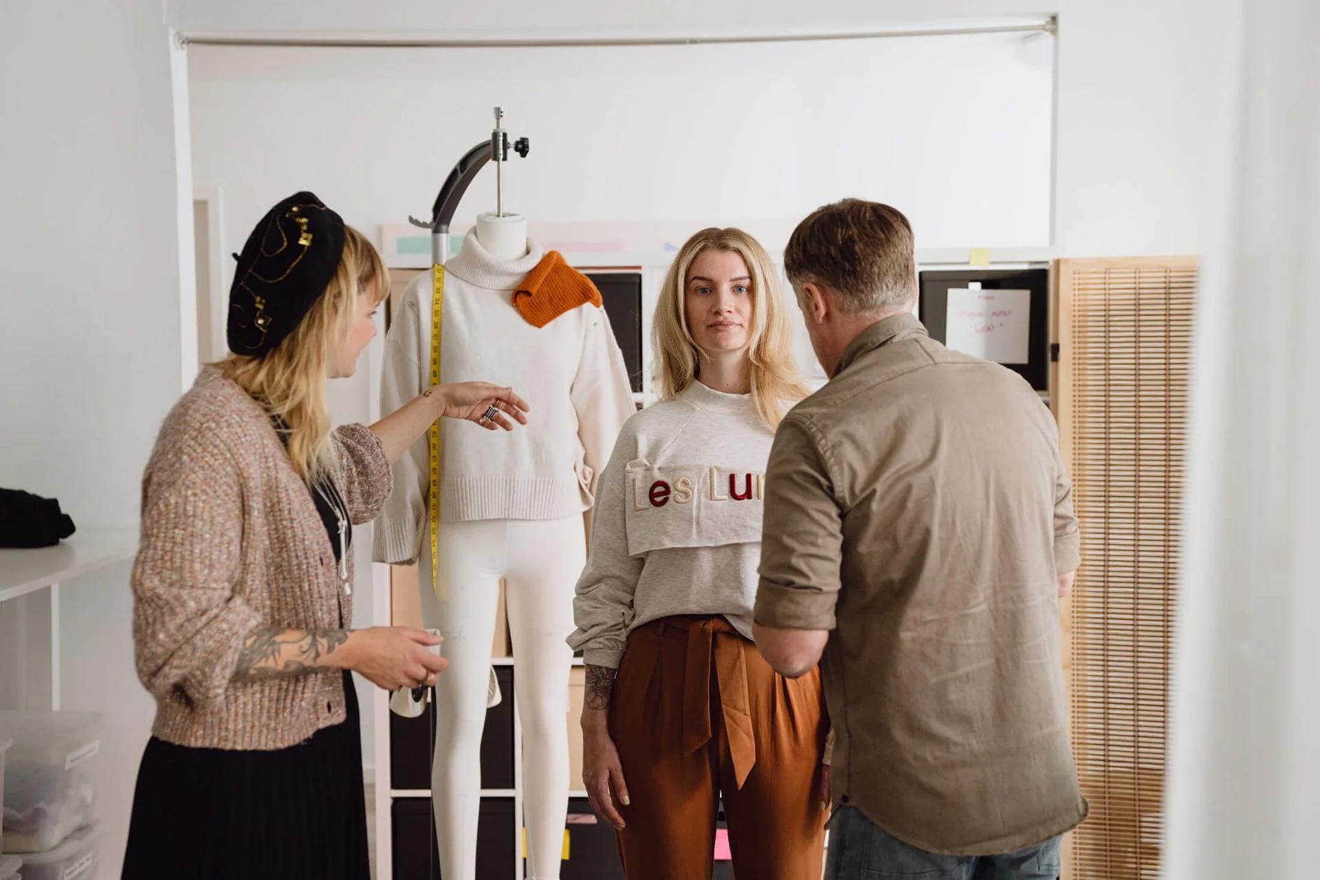 Drei Menschen sprechen in einem Raum, in dem eine Modelpuppe mit weißen Kleidungsstücken steht. Die Frau in der Mitte trägt eine graue Sweatshirt mit buntem Schriftzug und braune Hosen, die man in der Taille binden kann. Die Frau auf der linken Seite trägt einen türkisen Hut und einen Cardigan, während der Mann rechts eine beige Jacke trägt.