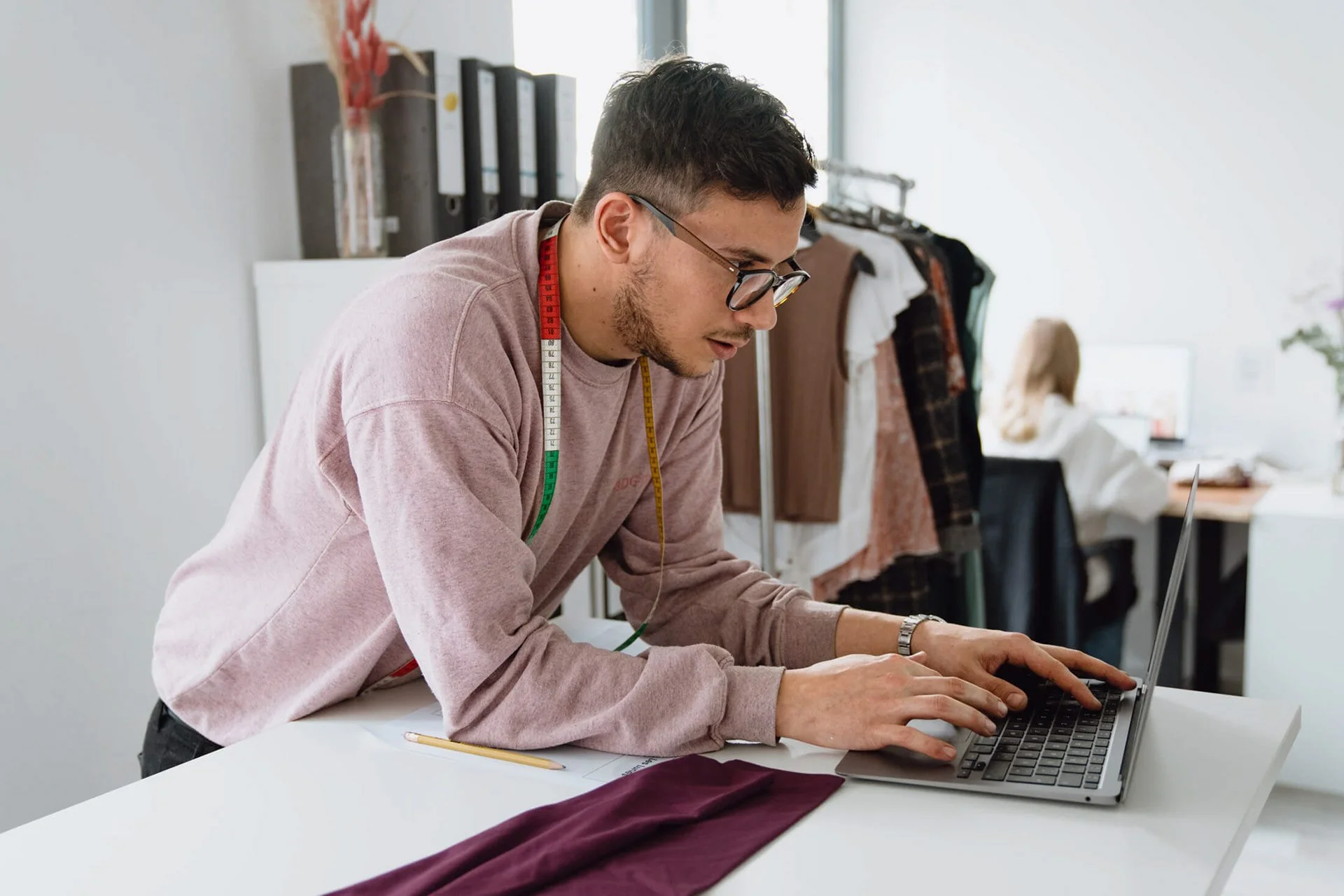 Mann arbeitet auf einem Laptop in einem Modeatelier, mit Stoffen und Kleidungsstücken im Hintergrund.