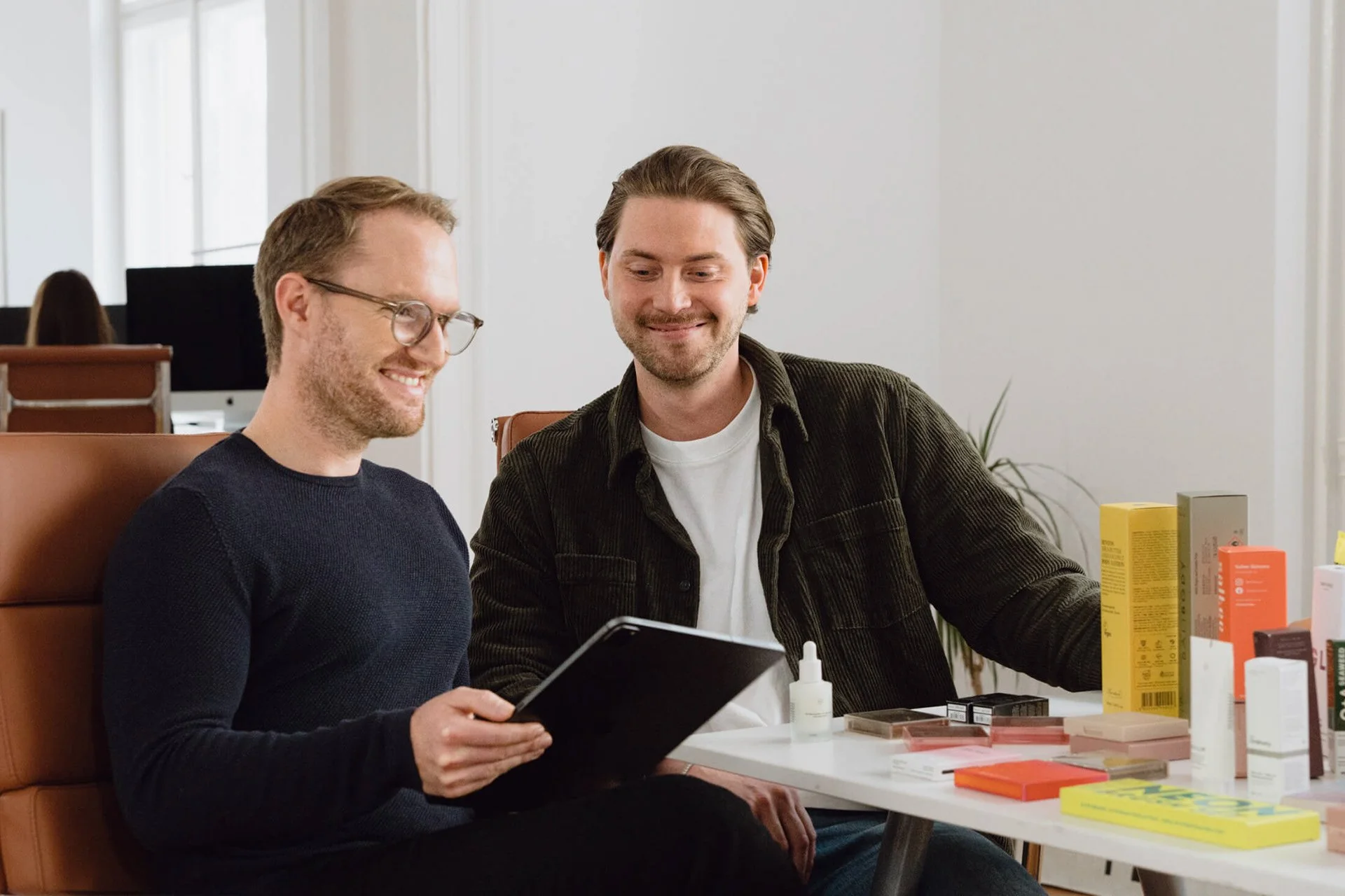 Zwei lachende Männer im Büro, die sich über ein Produktkatalog unterhalten, mit verschiedenen Wellness- und Kosmetikprodukten auf dem Tisch.