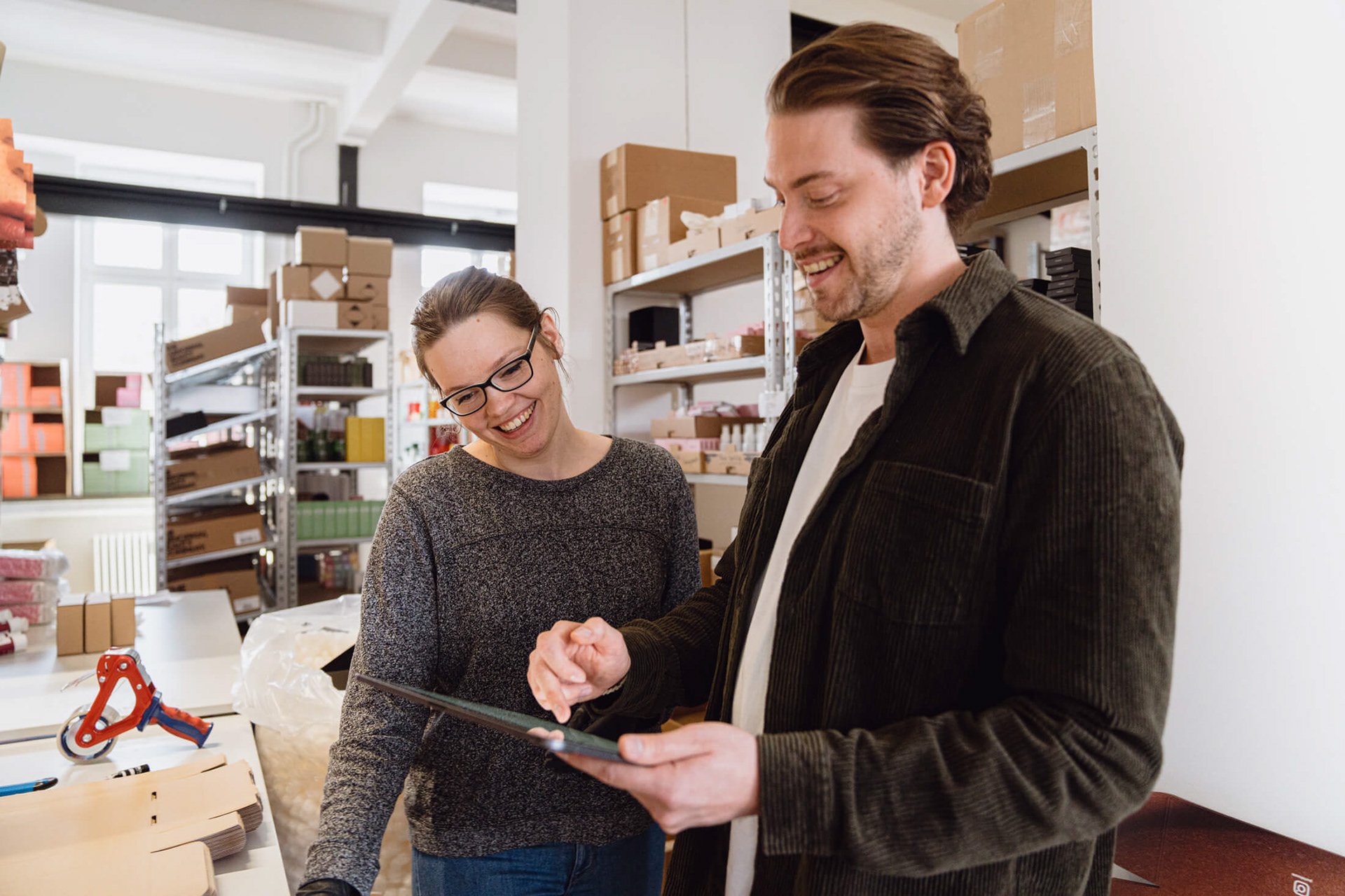 Zwei Menschen arbeiten in einem Lager, lachen und schauen auf ein Tablet, Regale im Hintergrund, Lagerutensilien auf Tischen.