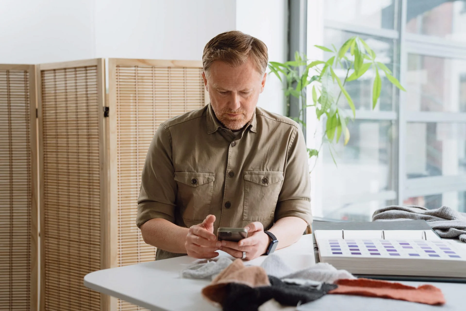 Mann sitzt an einem Tisch und schaut auf sein Smartphone, mit Stoffproben und Farbcharts vor ihm, im Hintergrund eine Zimmerpflanze.