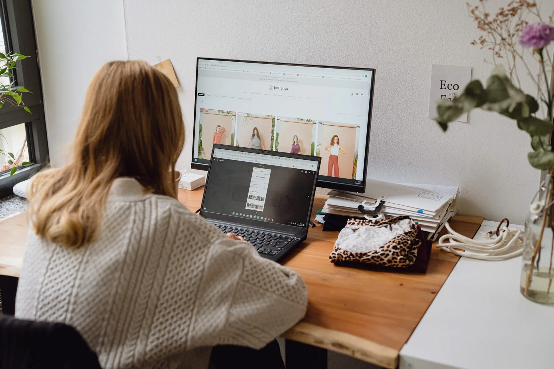 Eine Frau sitzt an einem Schreibtisch mit einem Laptop und einem großen Monitor, auf dem Modedesigns angezeigt werden. Der Schreibtisch ist mit Papieren, einem Stoffmuster, und anderen Büromaterialien übersät. Es ist eine helle, ordentlich eingerichtete Arbeitsumgebung mit Pflanzen und einem Fenster.