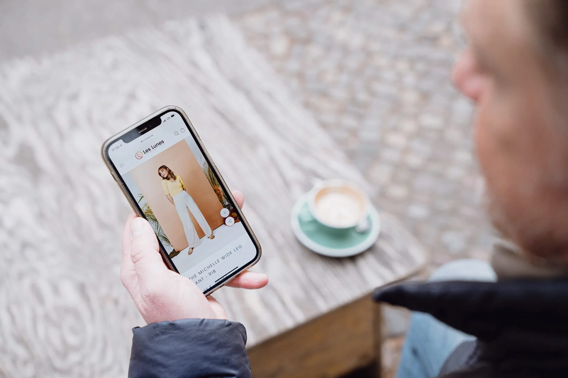 Nahaufnahme eines Mannes, der mit einem Smartphone ein Modefoto betrachtet, auf einem Holztisch steht eine Tasse Kaffee.