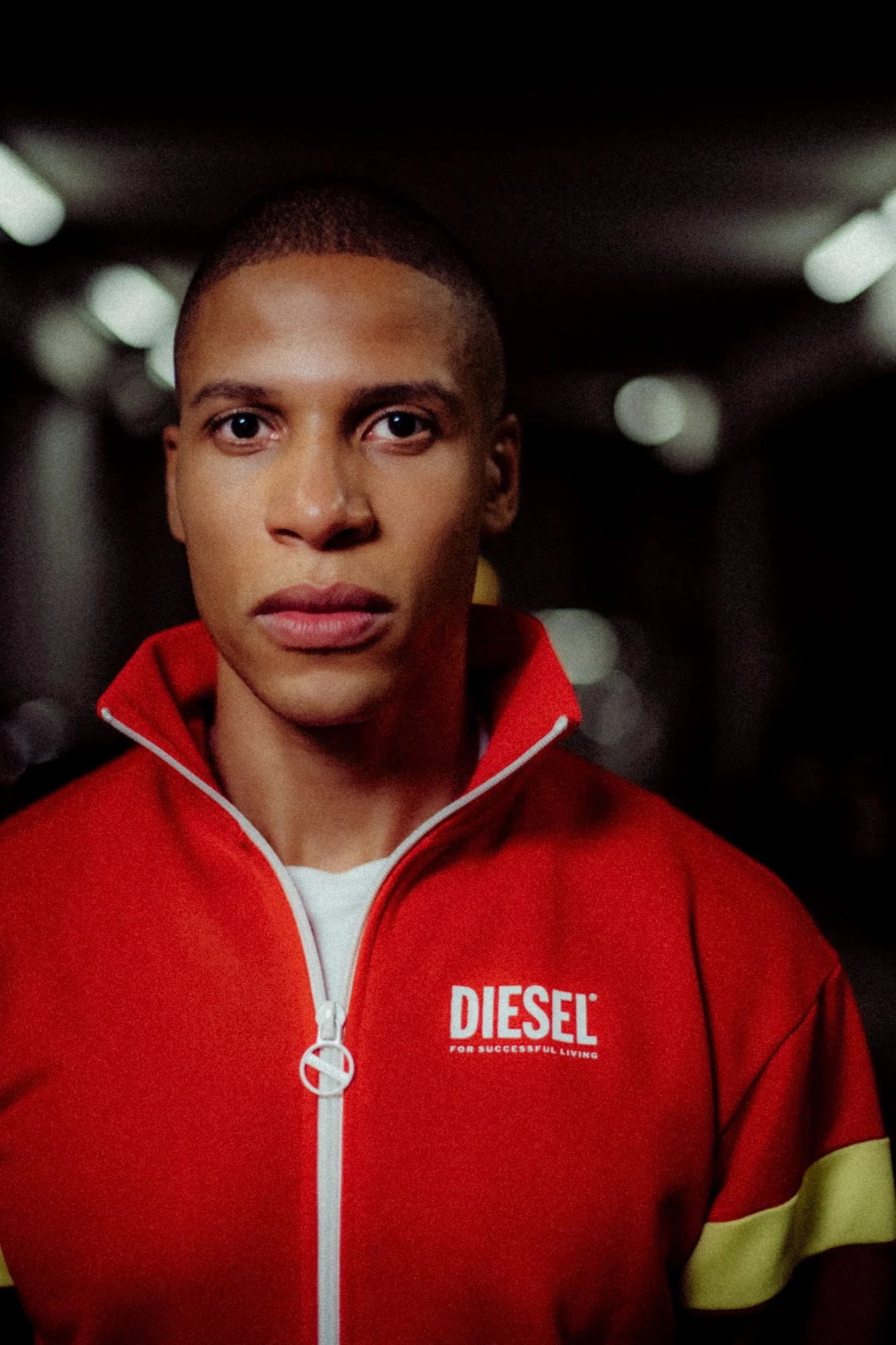 Ein junger Mann trägt einen roten Trainingsanzug mit dem Logo 'DIESEL' in einem dunklen Tunnel.