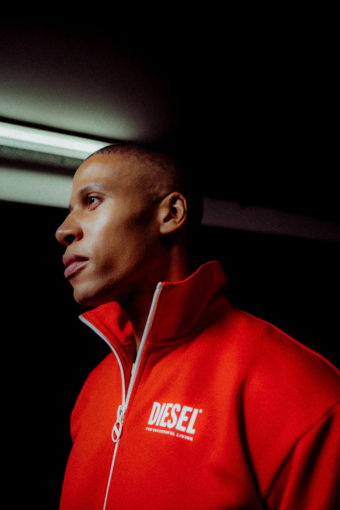 Nahaufnahme eines Mannes in einer roten Diesel-Jacke, der seitlich blickt, im dunklen Raum mit einer Lichtquelle über seinem Kopf.