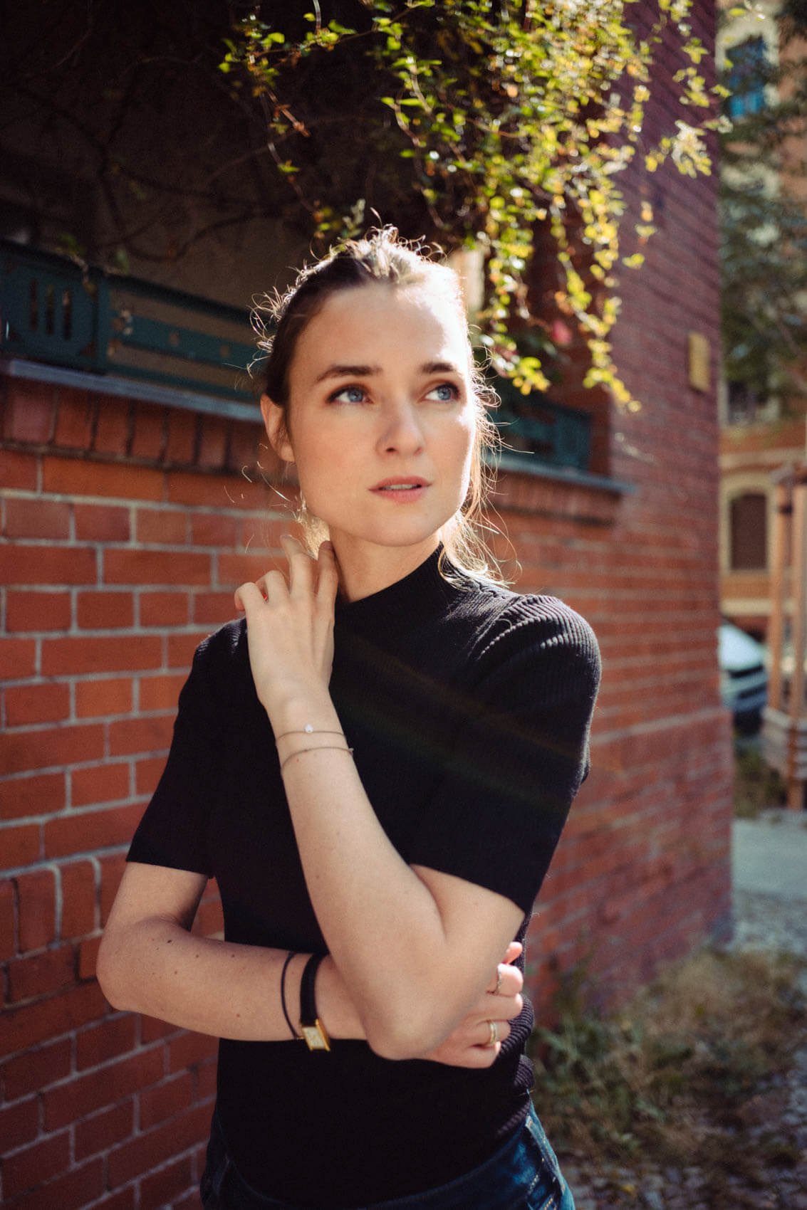 Josephin Busch mit langen Haaren, dunklem T-Shirt, steht vor einer Backsteinmauer im Sonnenlicht und schaut nach oben, umgeben von sonnigem Tageslicht und grünen Pflanzen.
