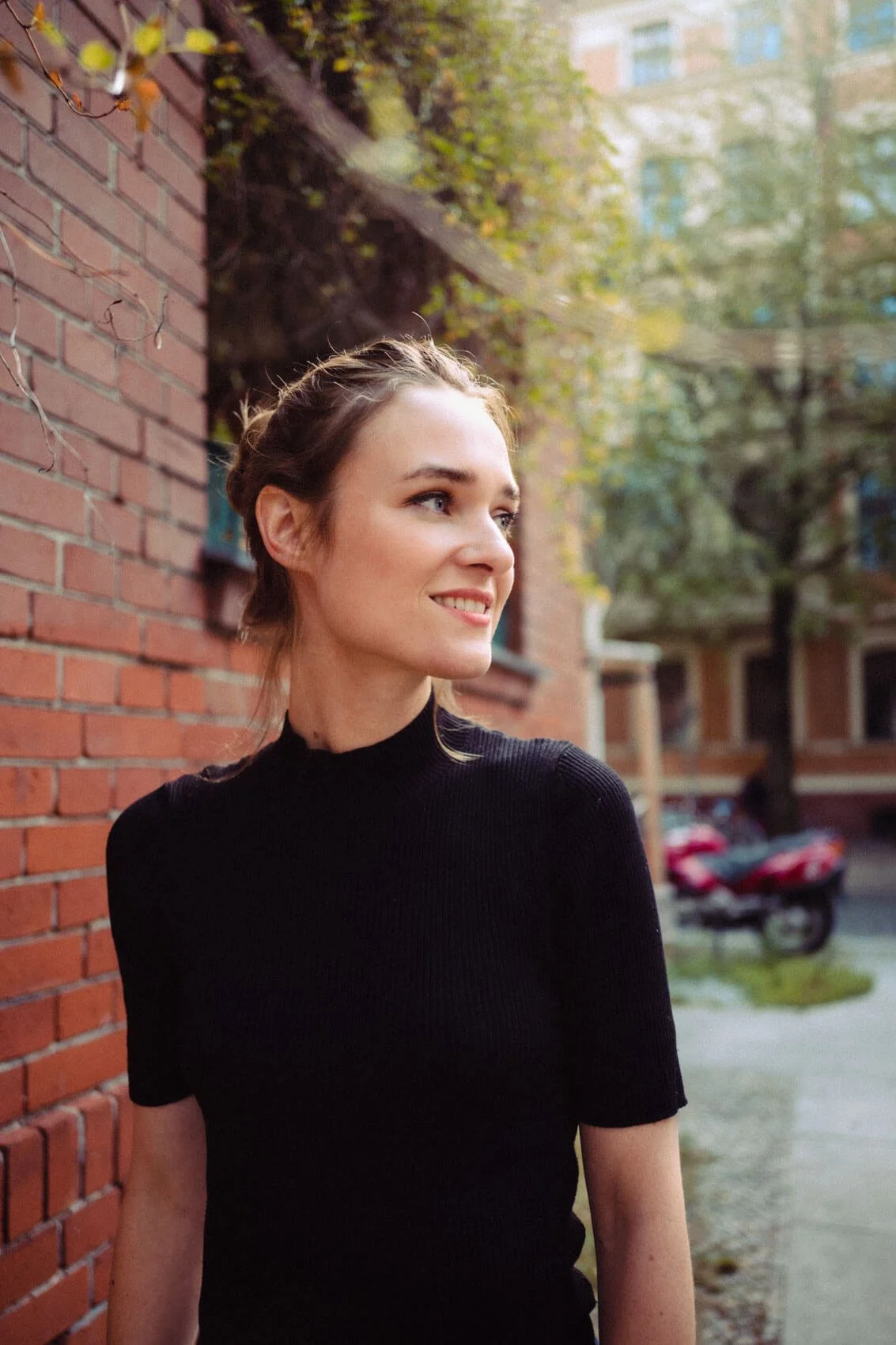 Josephin Busch mit braunen Haaren, gepinnten Haaren, schwarze Oberbekleidung, steht an einer Backsteinmauer im Freien, lächelt, im Hintergrund Bäume, Gebäude und ein geparktes Motorrad.