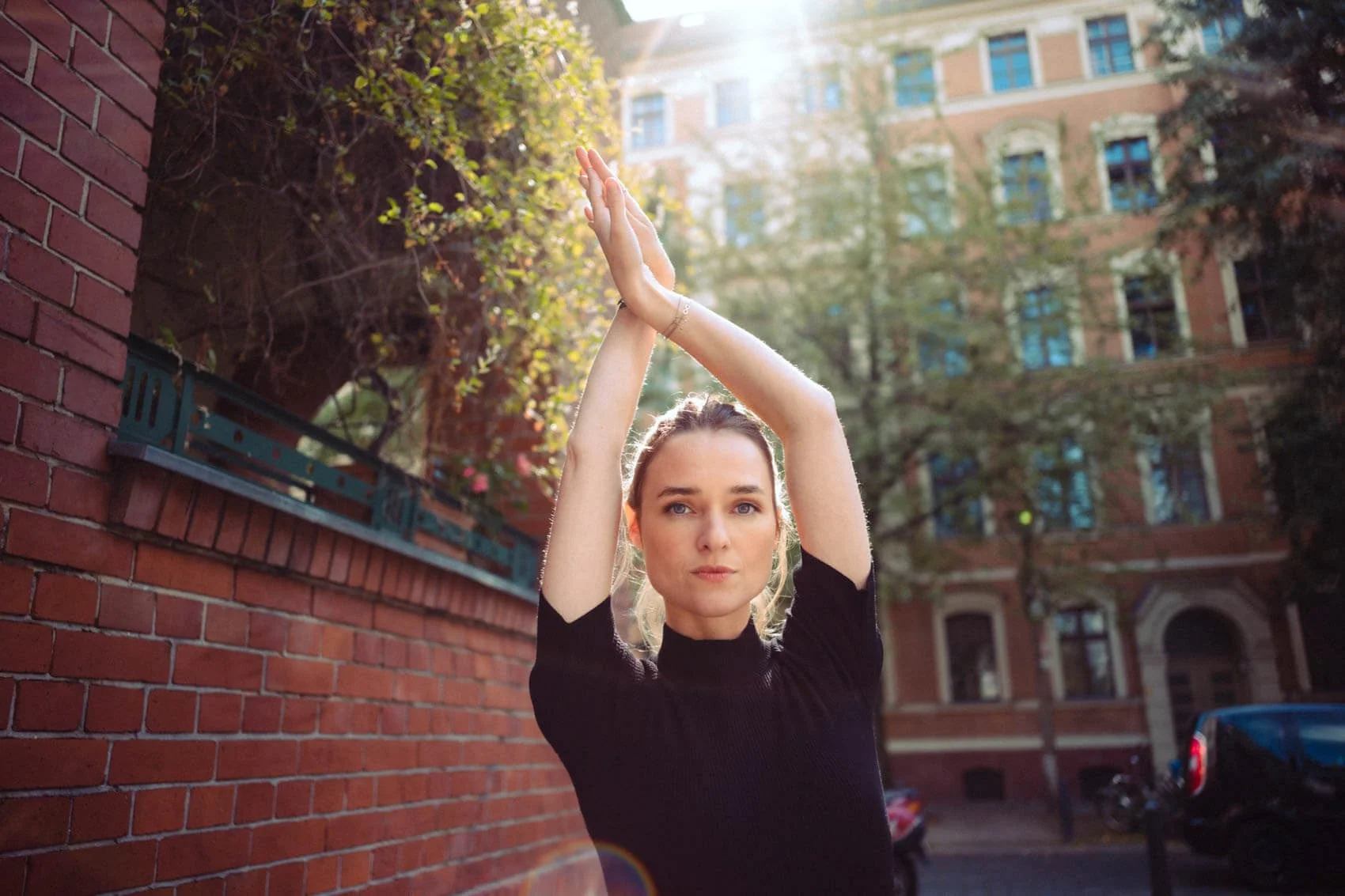 Josephin Busch in Schwarz, die draußen vor einem roten Backstein-Gebäude steht, die Hände über dem Kopf verschränkt, Sonnenlicht und Bäume im Hintergrund.