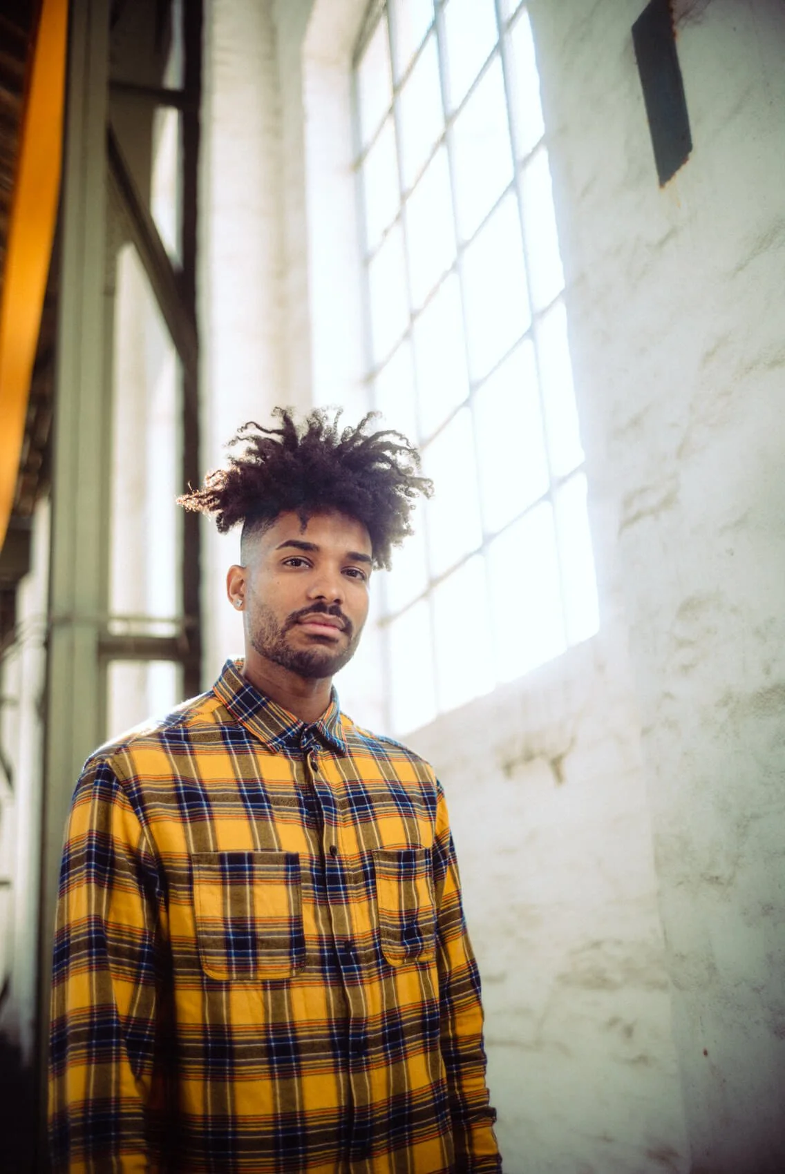Junger Mann mit dunklem Haarschopf und Bart trägt ein gelb-kariertes Hemd, steht vor einem großen Glasfenster in einem Industriegebäude.